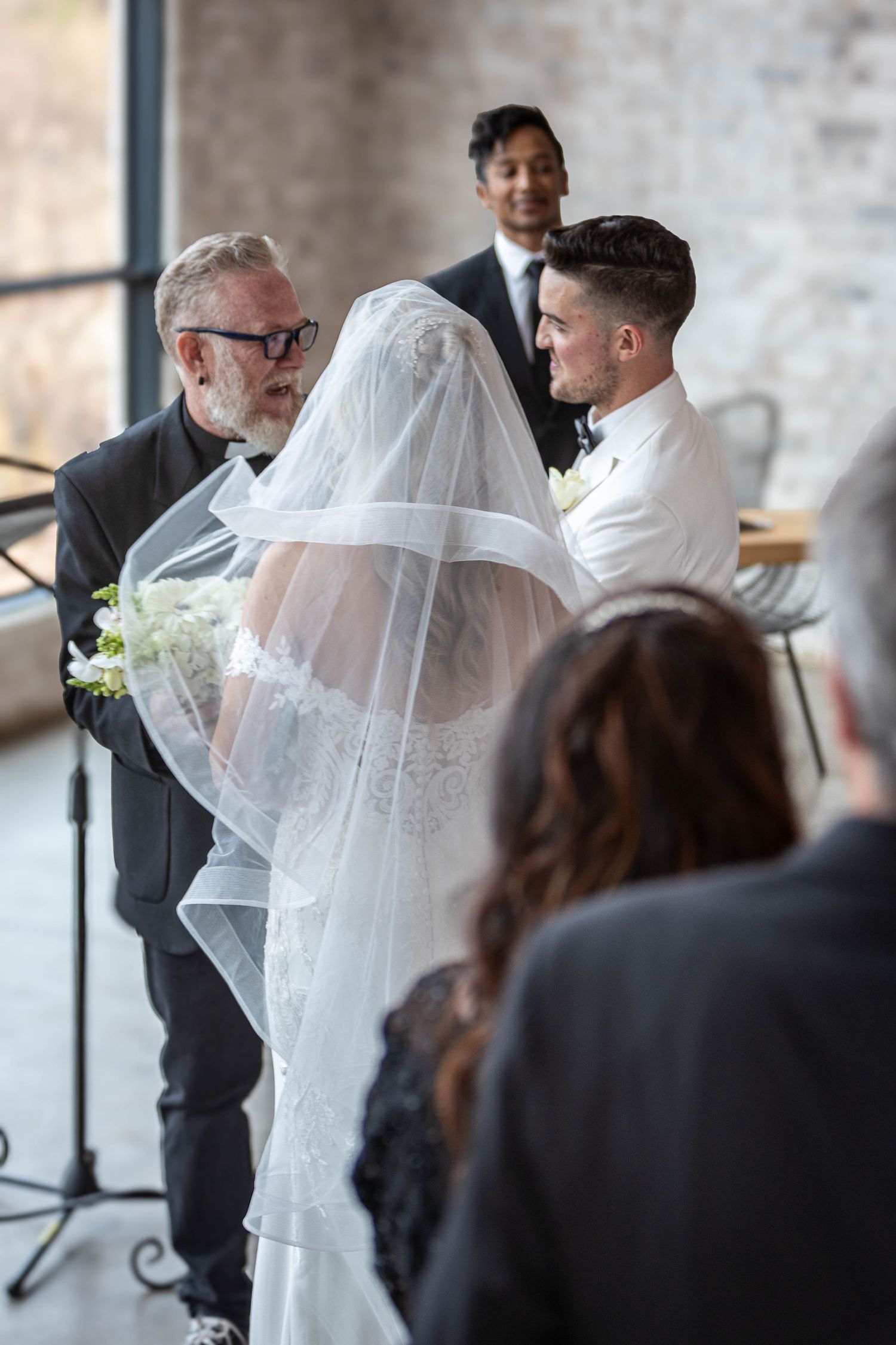 An officiant lifts the bride's veil as the groom watches lovingly during the indoor wedding ceremony.