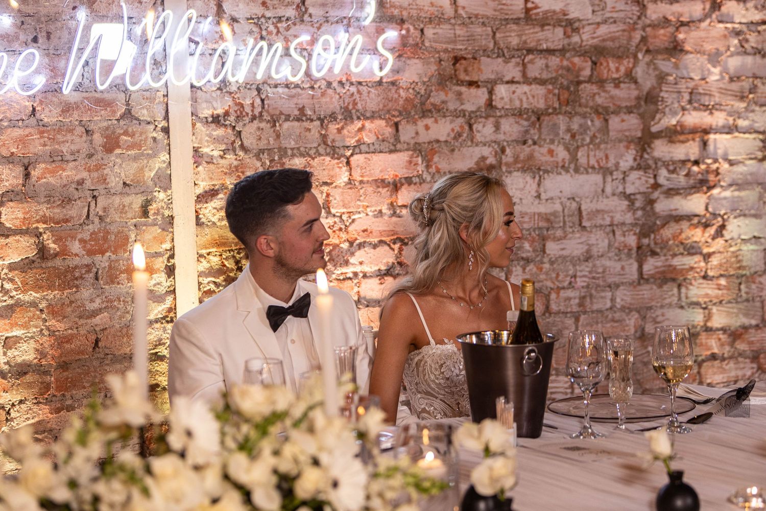 Bride and groom seated at sweetheart table beneath glowing Williamsons neon wedding sign.