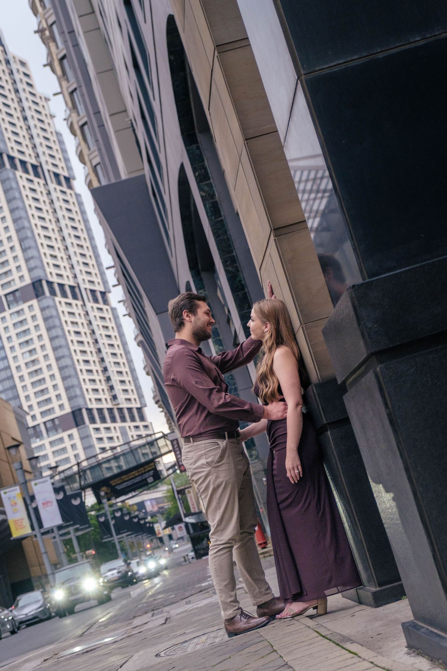 Couple embraces tenderly against modern glass skyscraper facade on city street in coordinating brown and purple outfits.