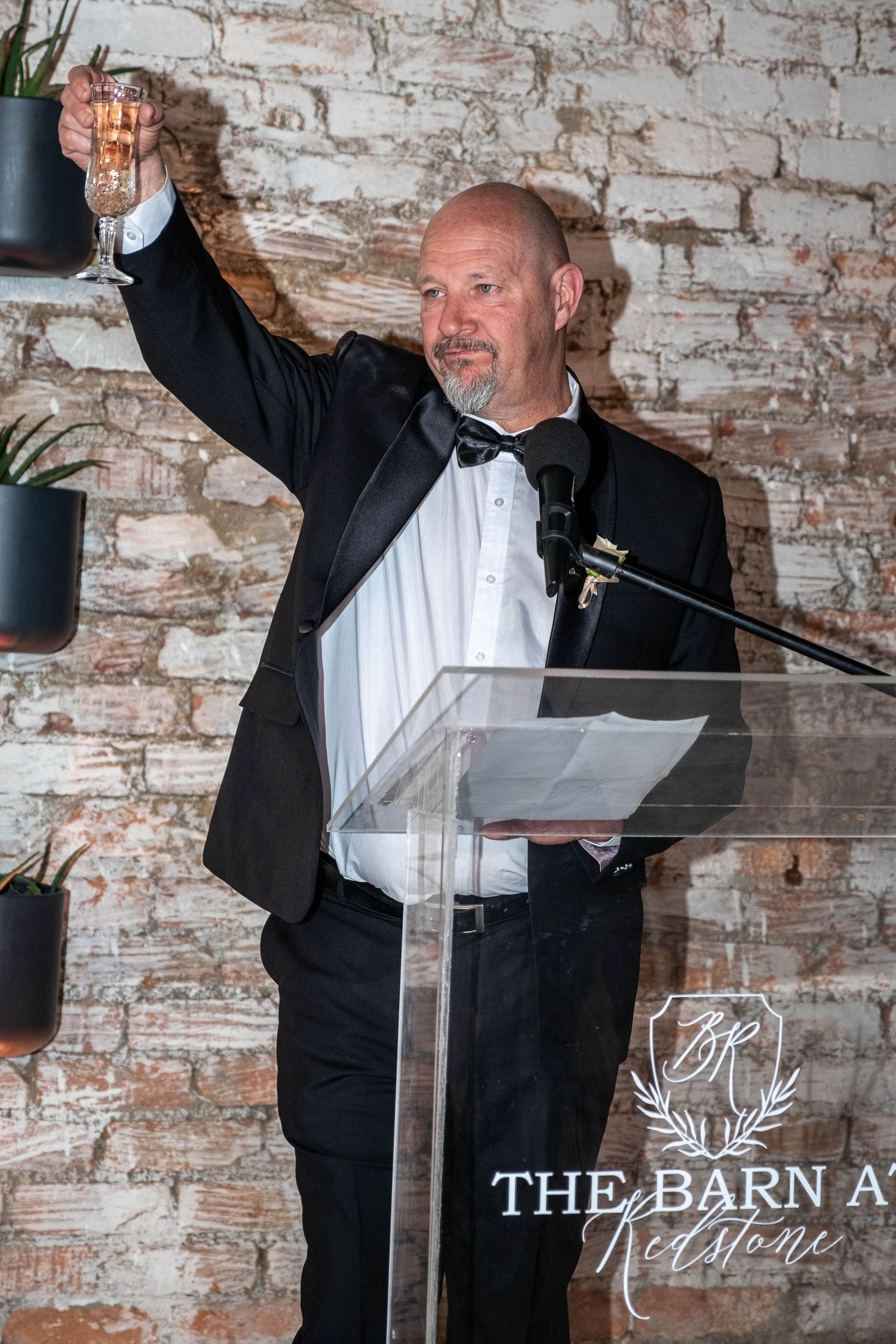 Man in tuxedo raises champagne glass in wedding toast at The Barn At Redstone venue.
