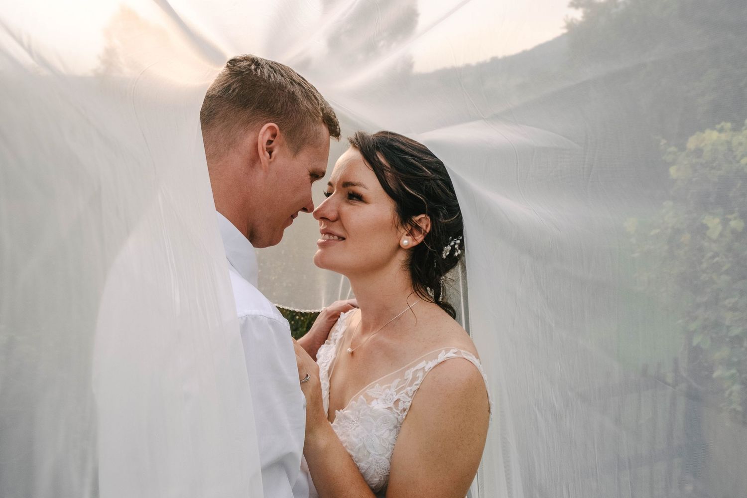 Bride and groom share an intimate forehead-to-forehead moment draped under a flowing white veil in soft golden light.