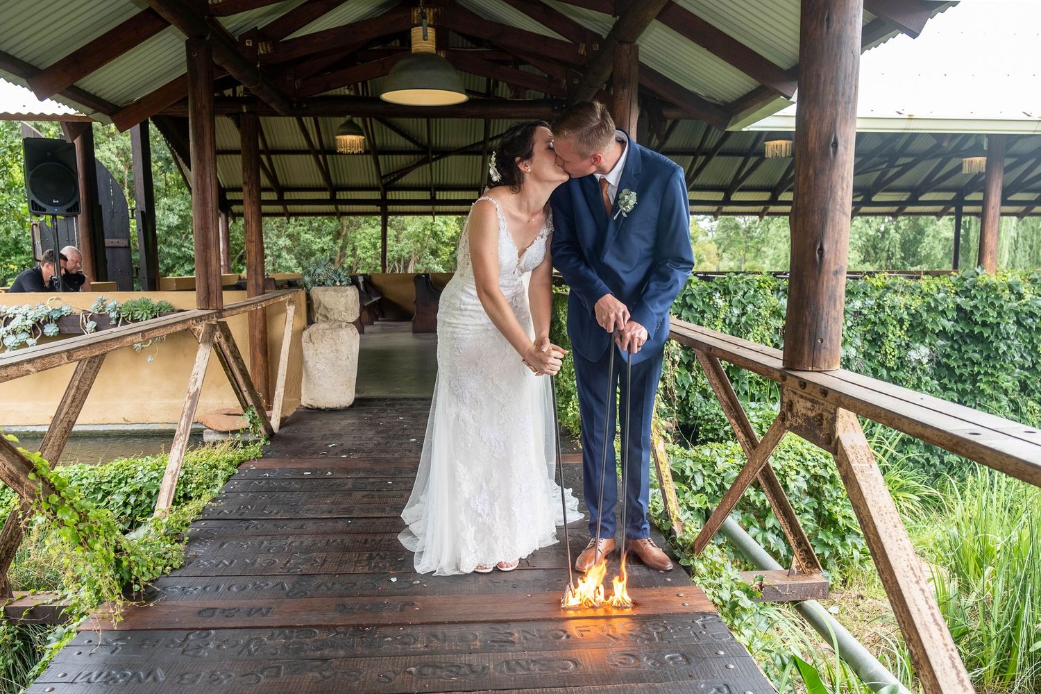 A newlywed couple performs a unity candle ceremony on a wooden deck surrounded by lush greenery.