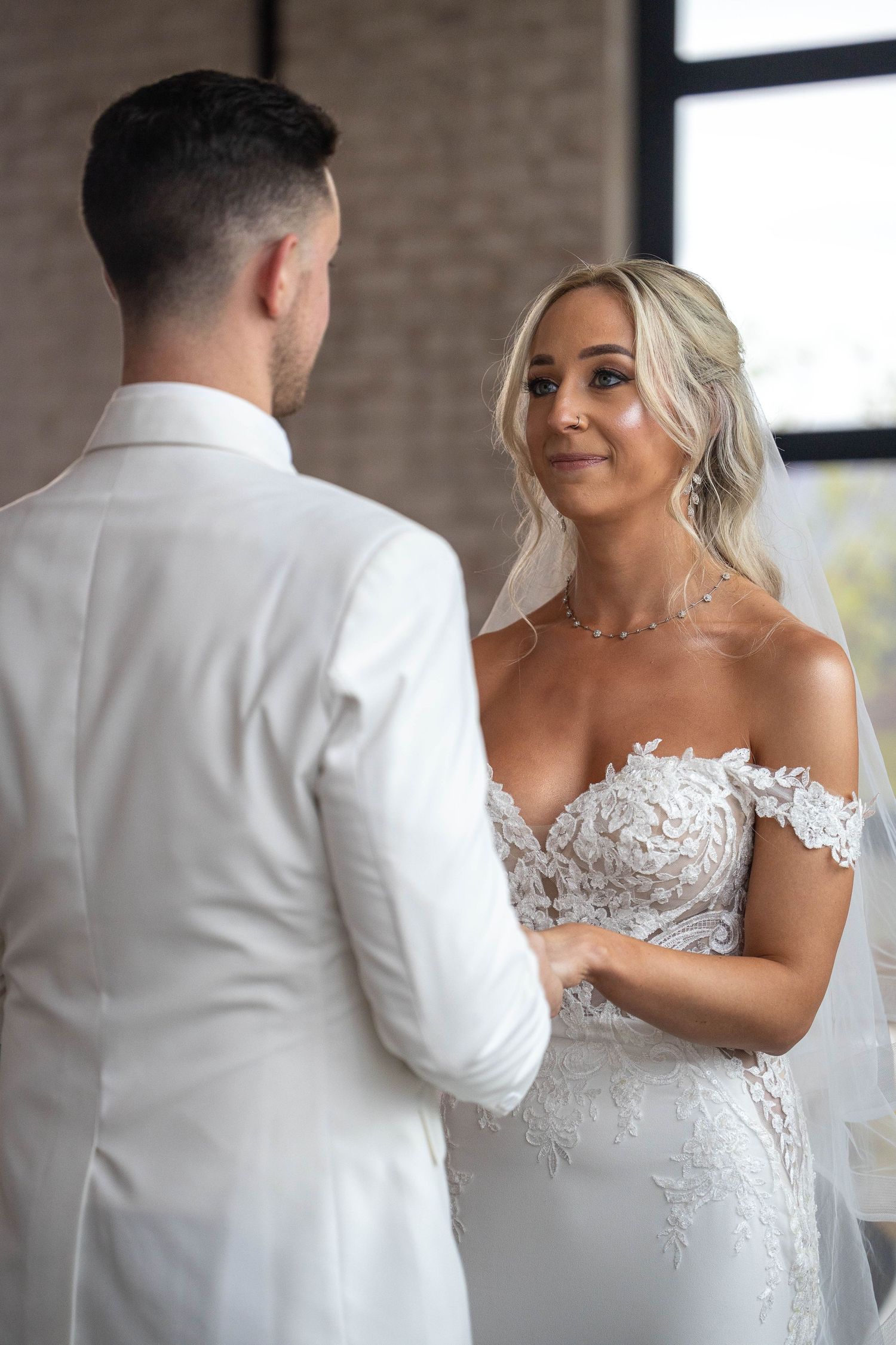 Bride in lace off-shoulder gown holds grooms hands during wedding ceremony.