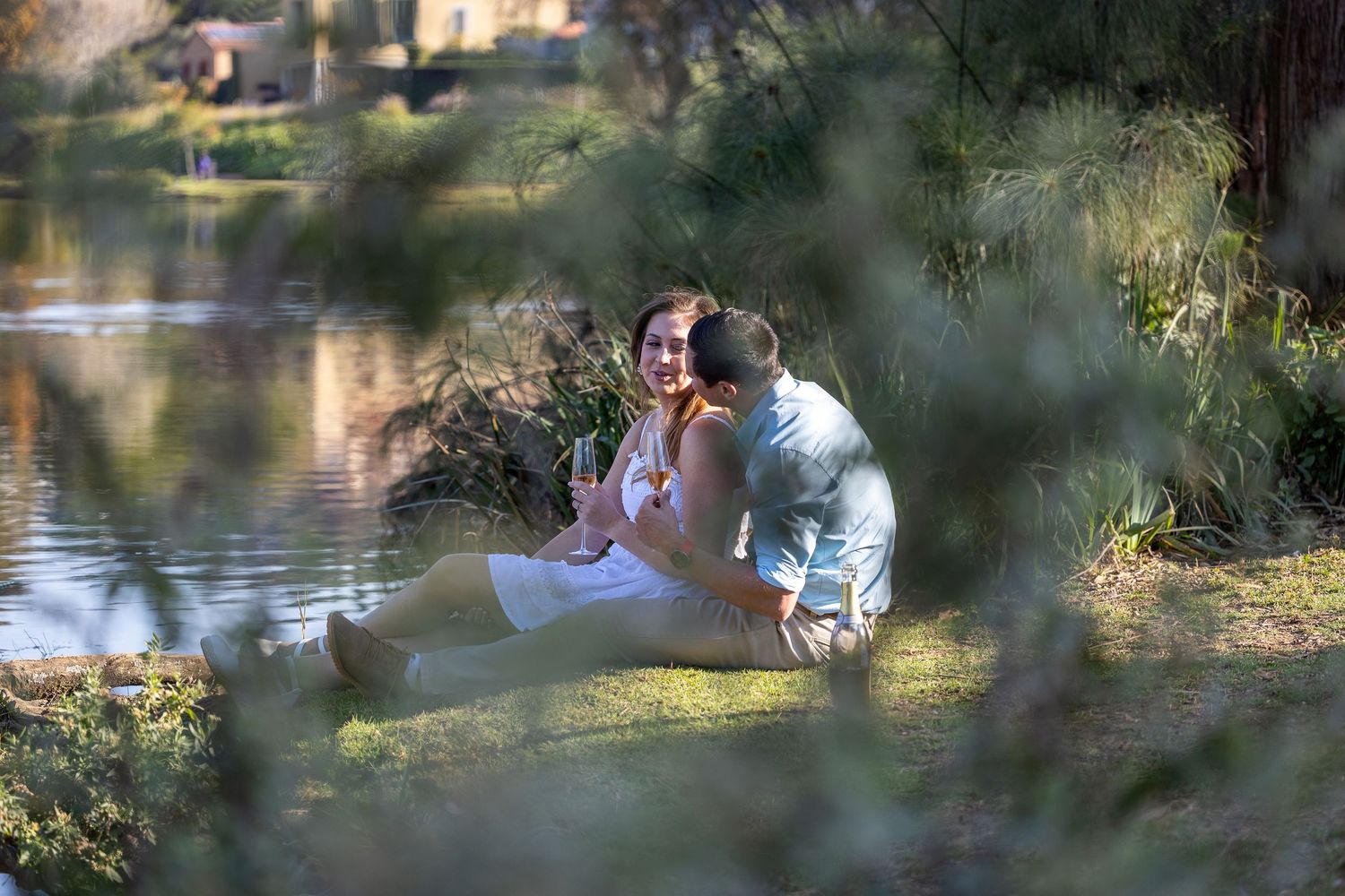 A couple sits intimately by a reflective pond, framed by soft foliage.