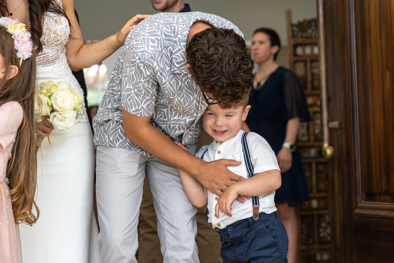 A man in a patterned shirt hugs a young boy in suspenders at a wedding celebration indoors.