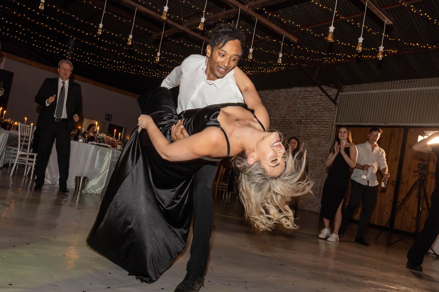 Couple performing dramatic dance dip on wedding reception dance floor with guests watching.