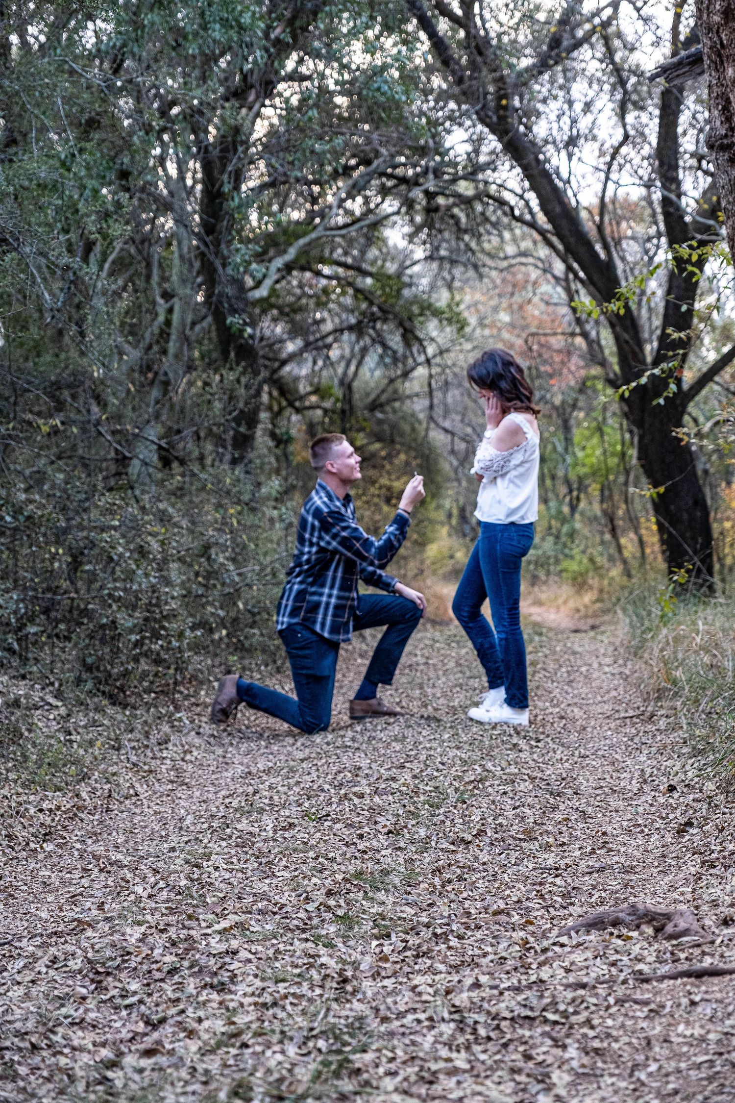 Man down on one knee proposing to his girlfriend on a leaf-covered woodland path during engagement shoot.