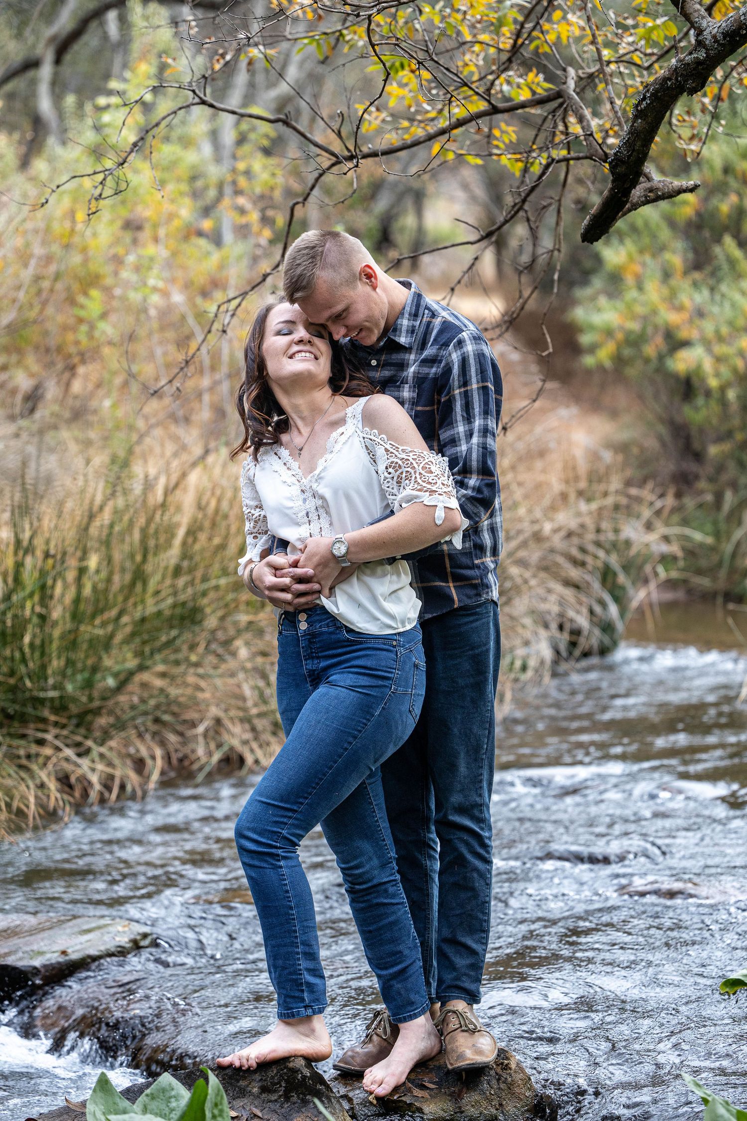 Romantic couple standing barefoot on rocks in a creek, man embracing woman from behind in autumn setting.