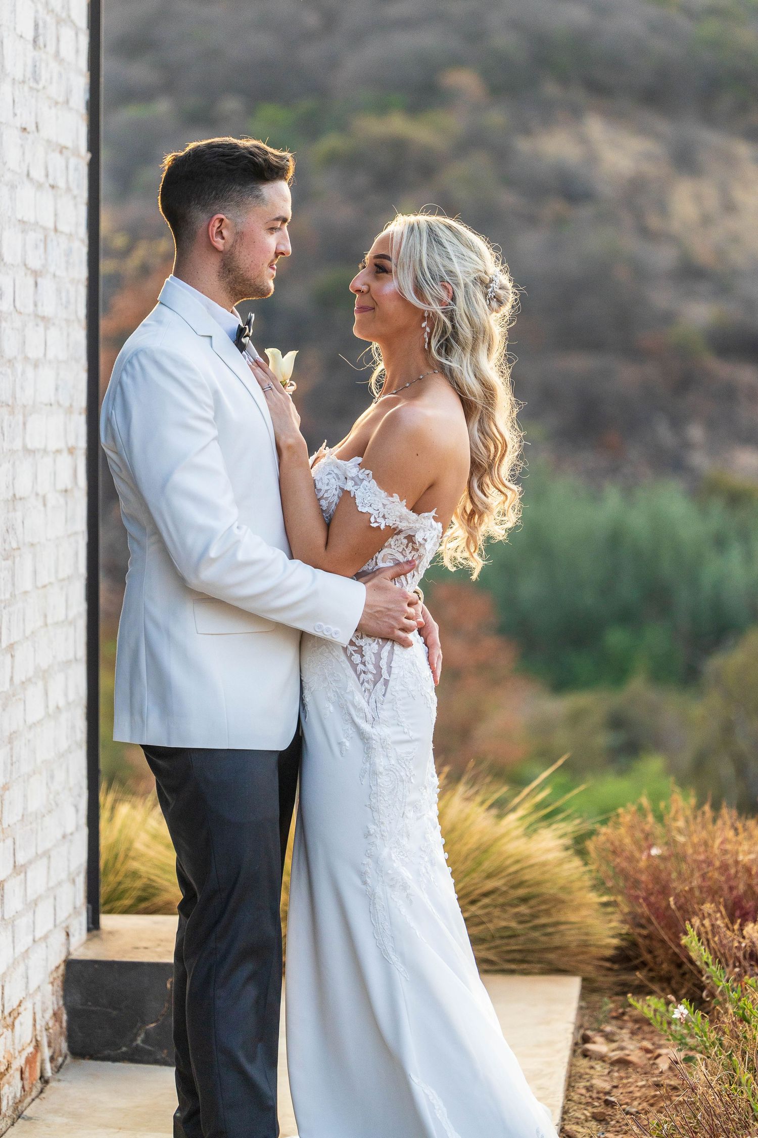 Bride and groom in white wedding attire embrace romantically outdoors with golden hillside scenery behind them.