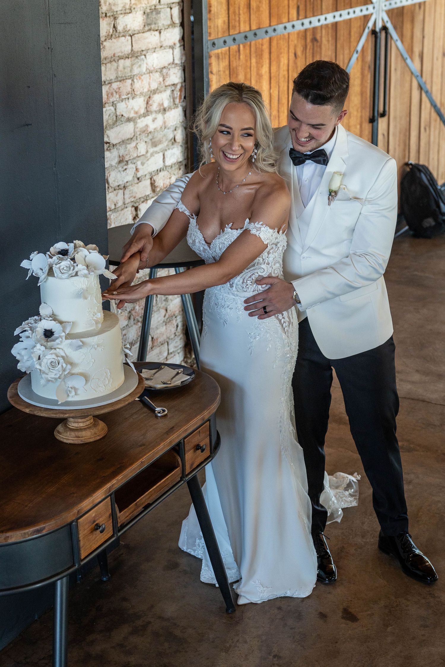 Happy bride and groom cutting white floral wedding cake together at rustic barn wedding reception.