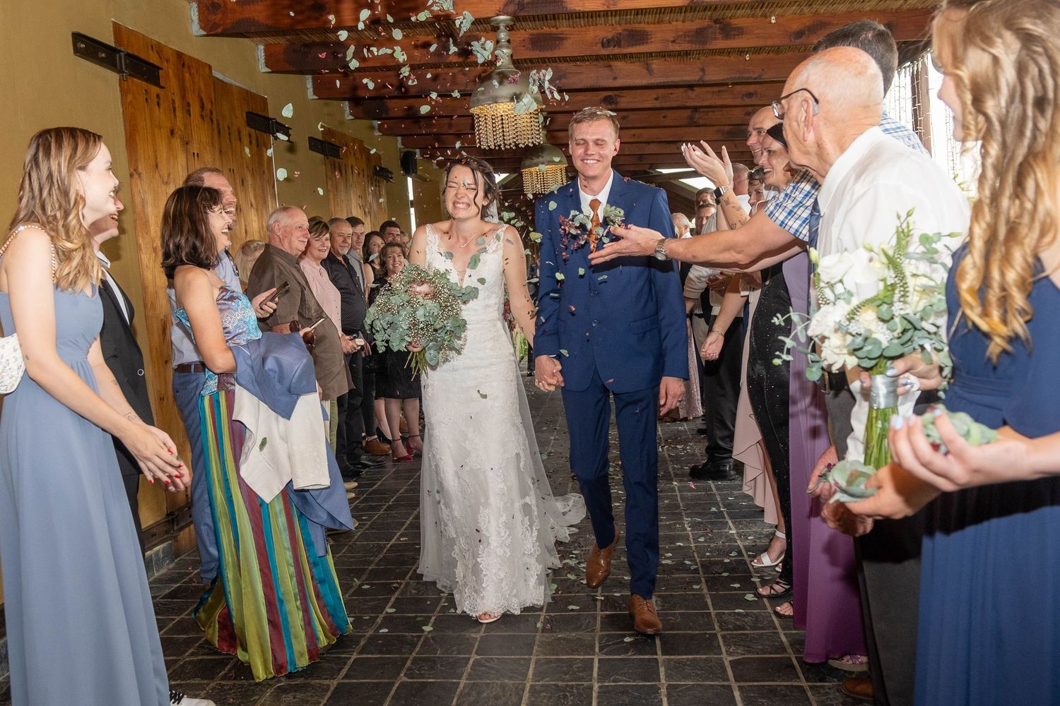 Smiling newlyweds walk down the aisle as guests toss confetti in a rustic indoor wedding venue.