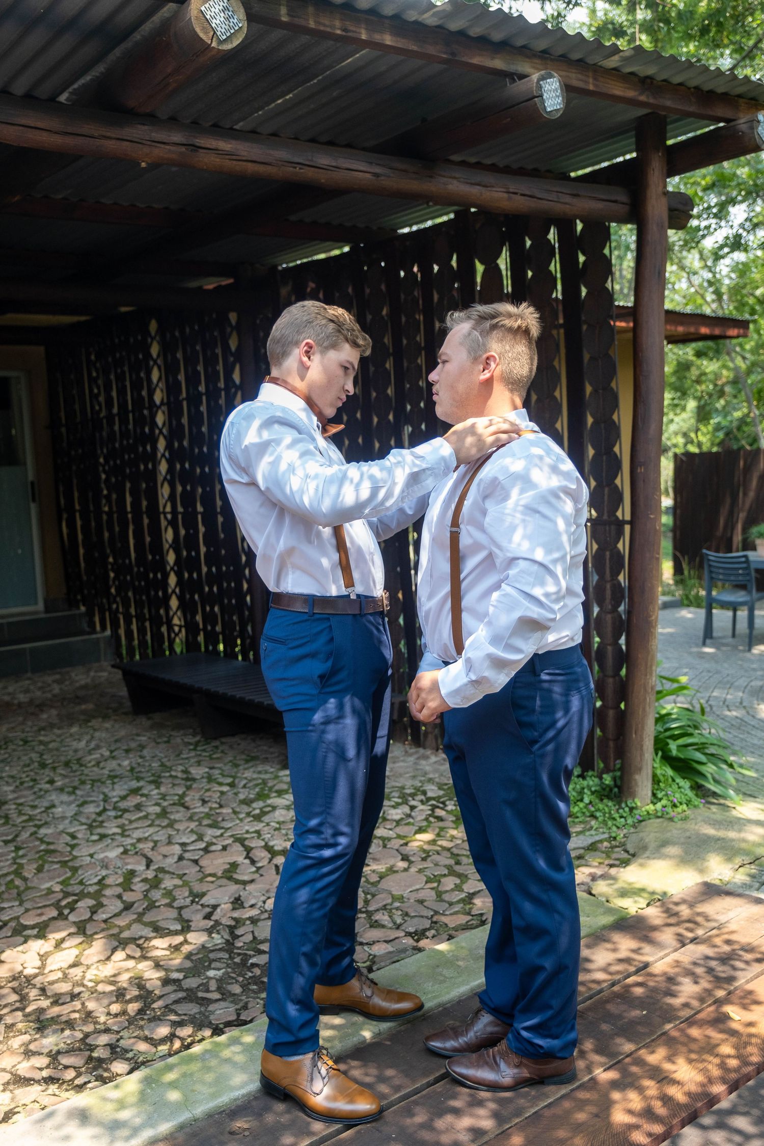 Two groomsmen in white shirts and blue pants with brown suspenders help each other prepare for a wedding outdoors.