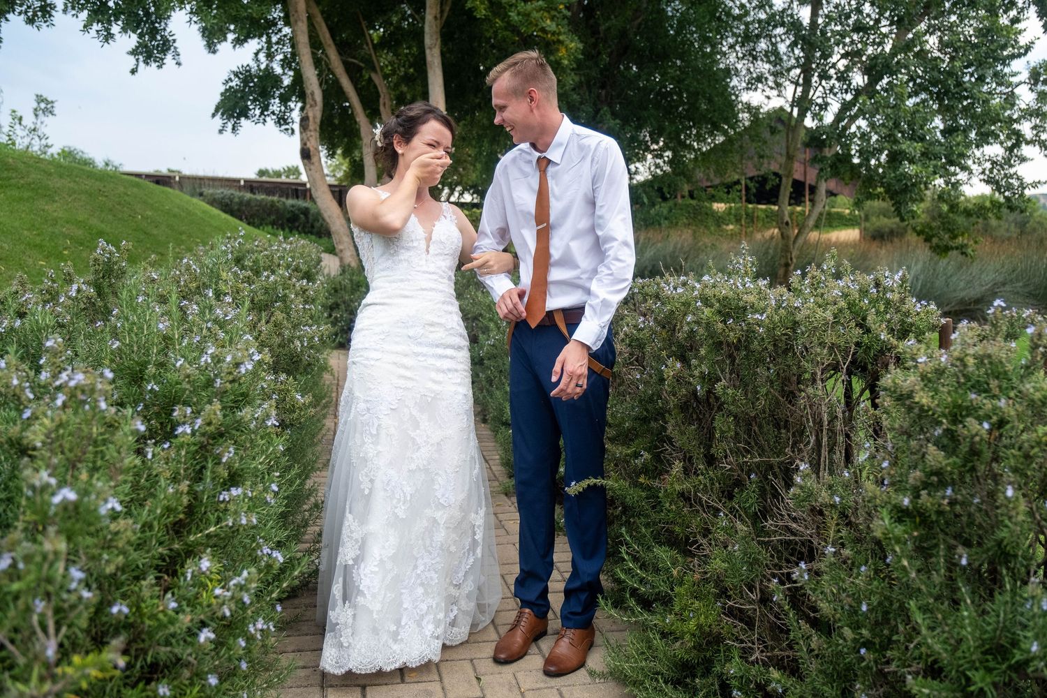 Bride laughs while covering her mouth as groom smiles beside her on a garden path lined with purple flowering shrubs.