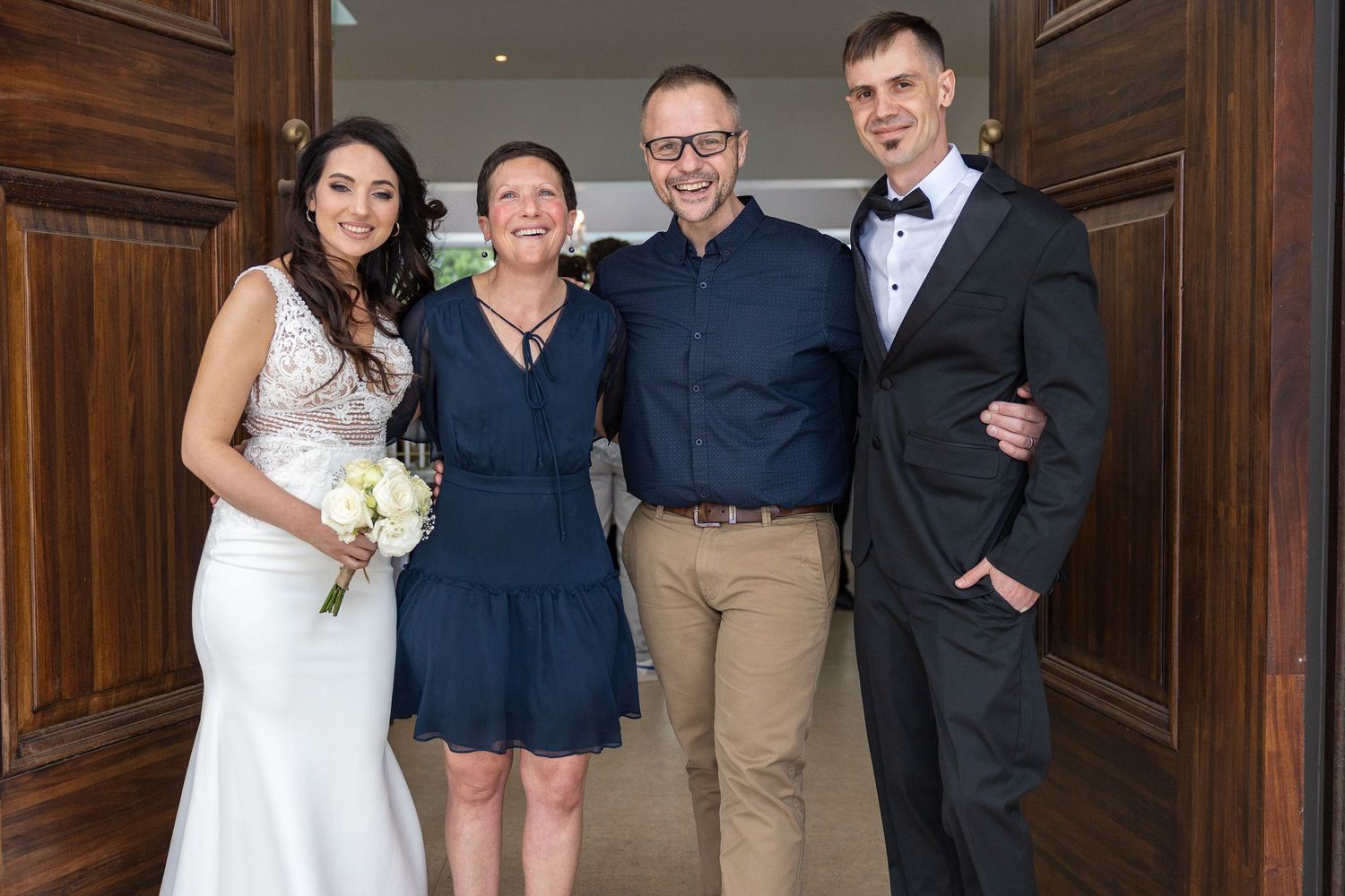 A bride holding a bouquet poses with three guests in front of wooden doors at a wedding venue.