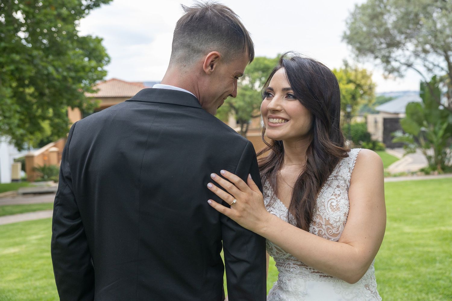 Smiling bride in lace wedding dress places hand on groom's chest during outdoor wedding photos.