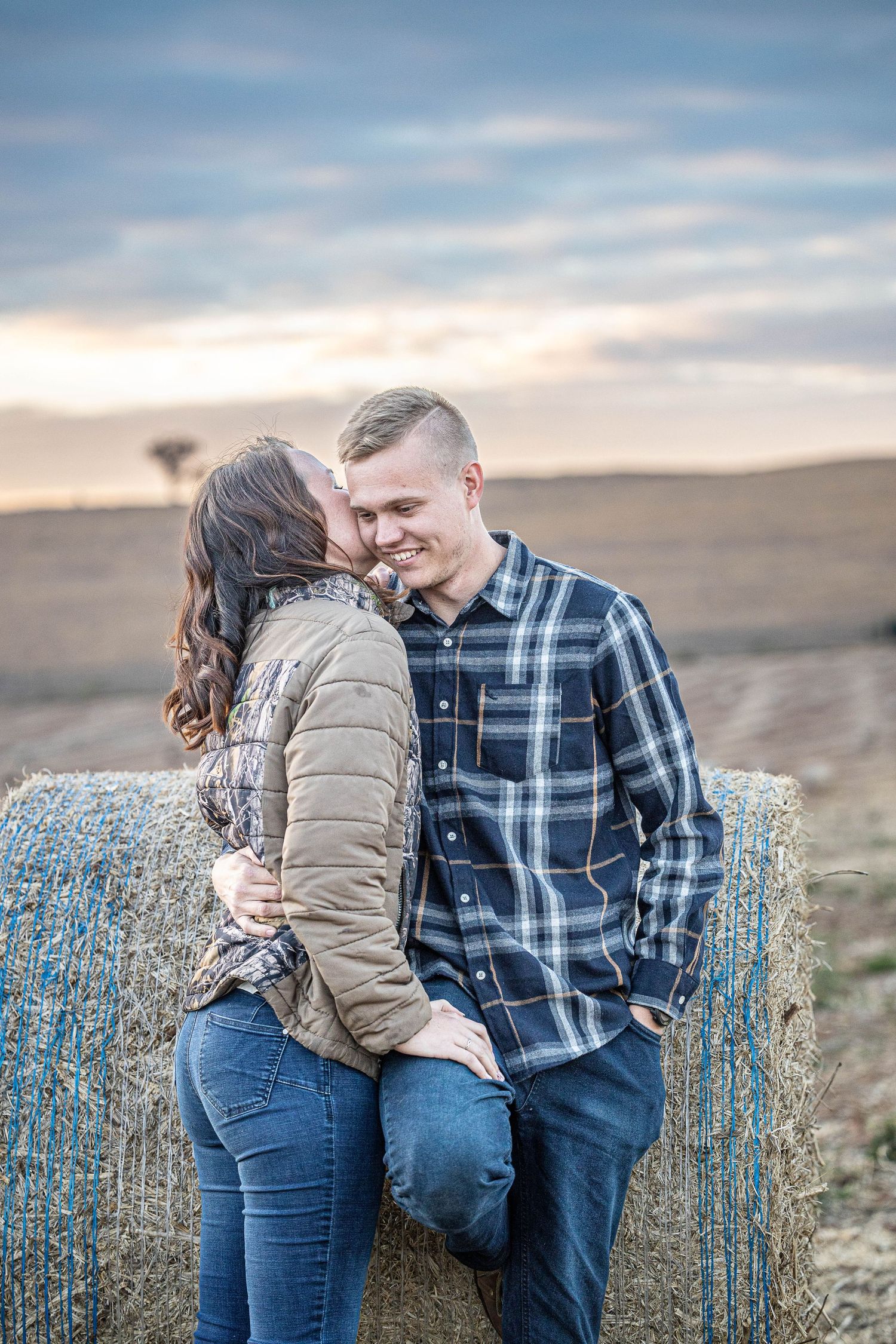Woman whispering to smiling man leaning on hay bale in golden field during romantic sunset session.