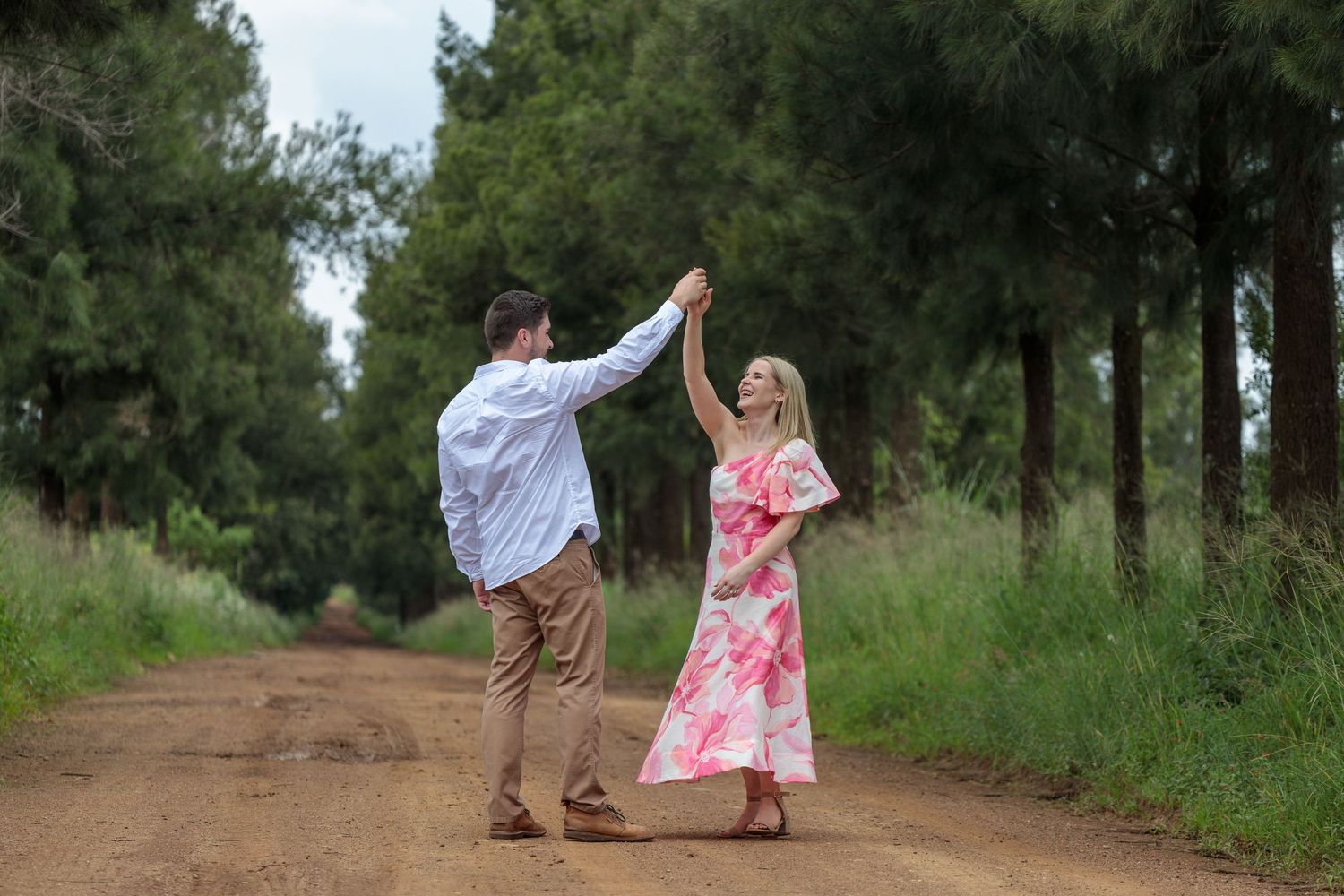Joyful couple dancing on a dirt road surrounded by tall green trees.