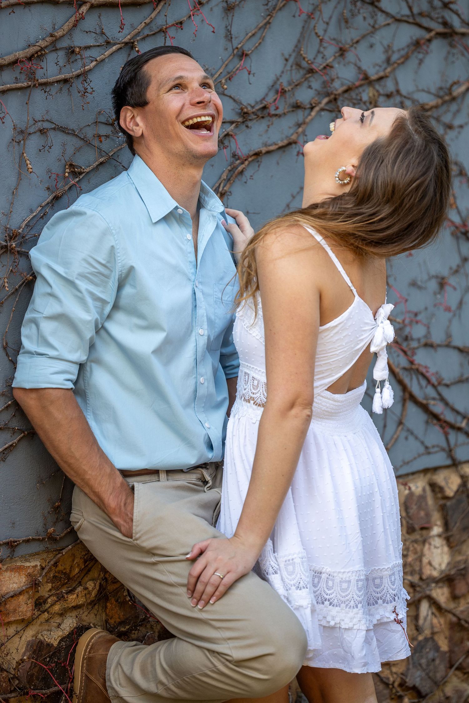Laughing couple posing against a vine-covered wall, woman in white dress touching man in light blue shirt.