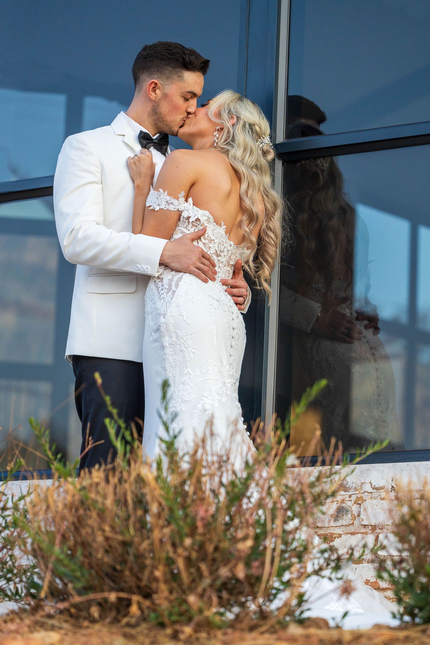 Newlyweds share a passionate kiss outside glass building, groom in white tuxedo, bride in lace mermaid gown.