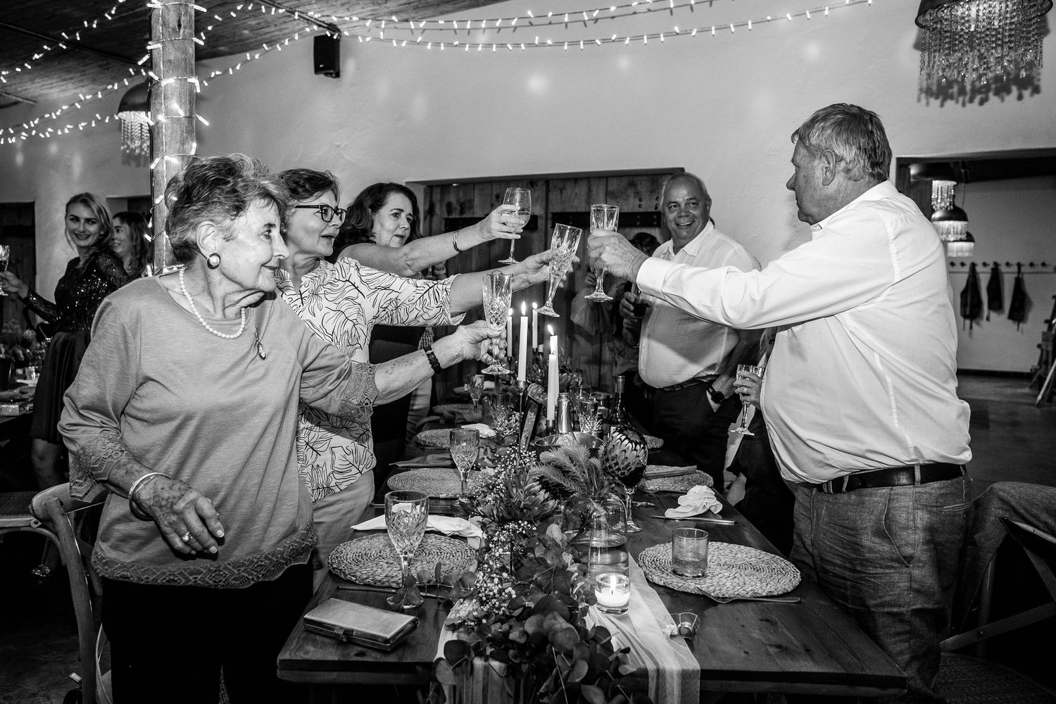 Wedding guests raise glasses in a celebratory toast around a decorated table at a rustic reception venue.