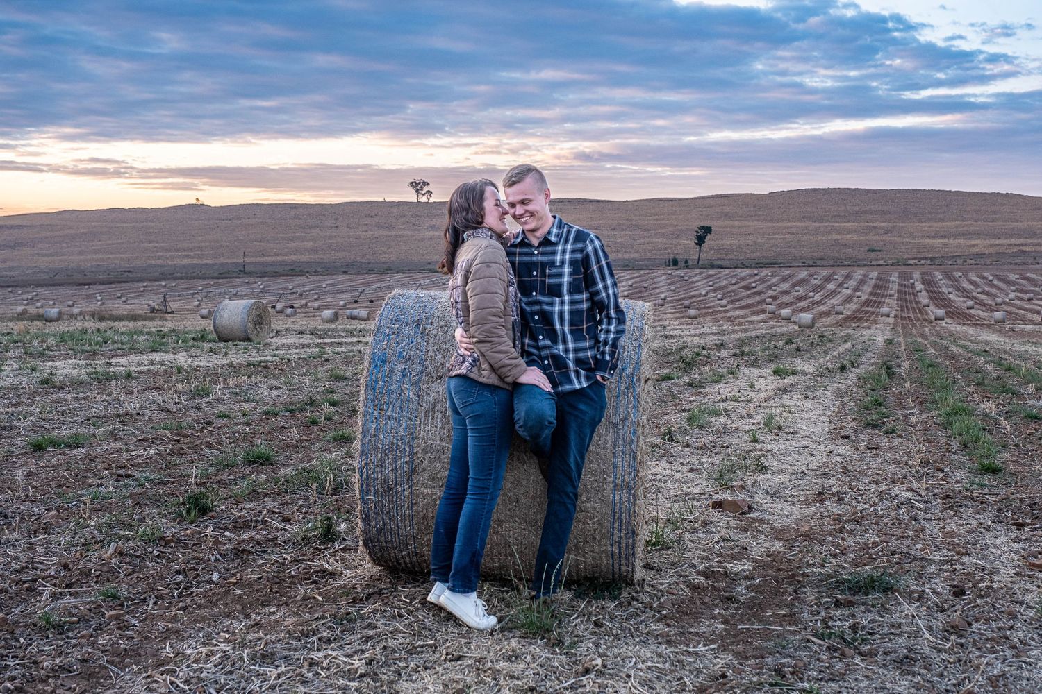 Couple sharing a kiss while leaning against round hay bale in wide open field at sunset.