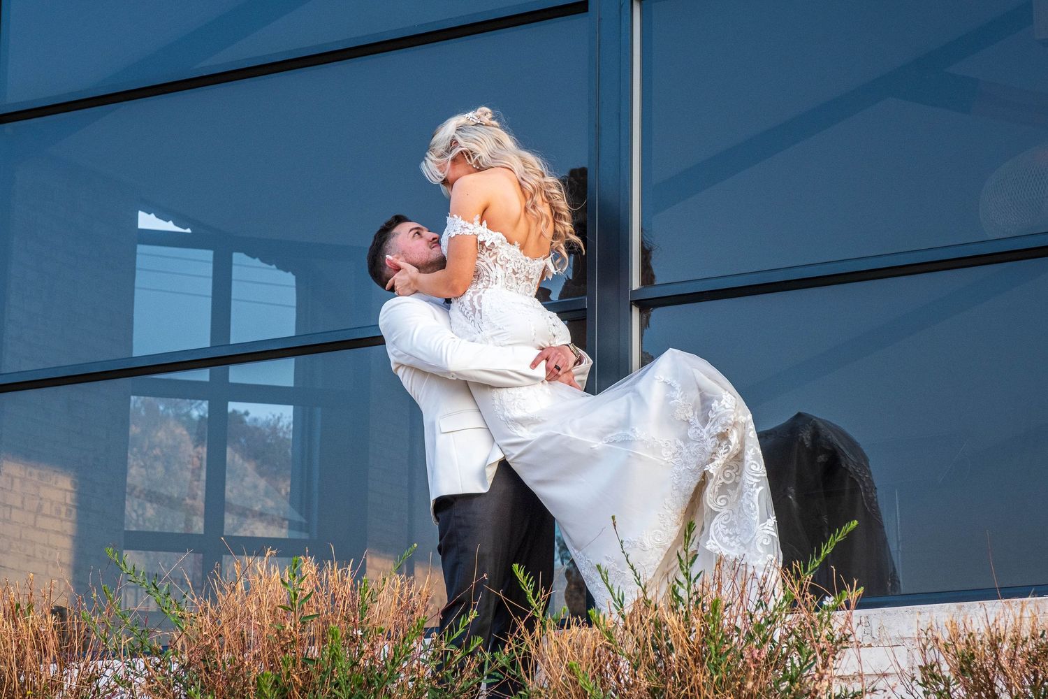 Groom lifts bride in white lace gown for a kiss in front of modern glass building exterior.