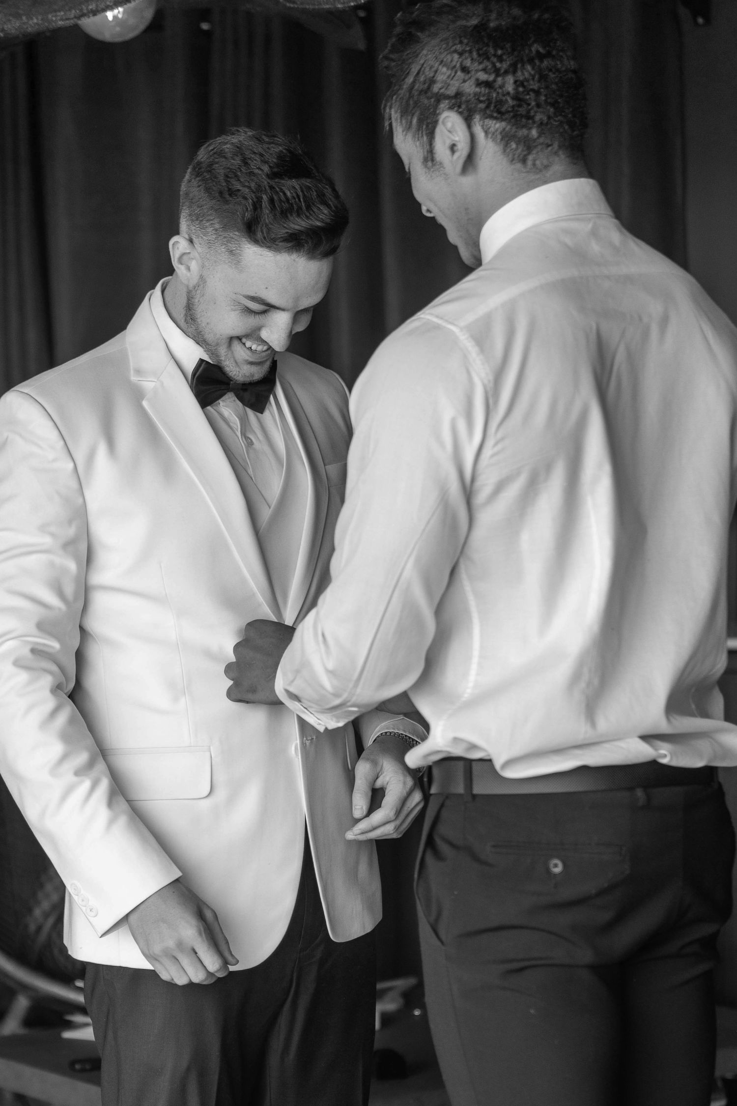 A groom in a white tuxedo jacket smiles as his groomsman helps adjust his suit before the wedding ceremony.