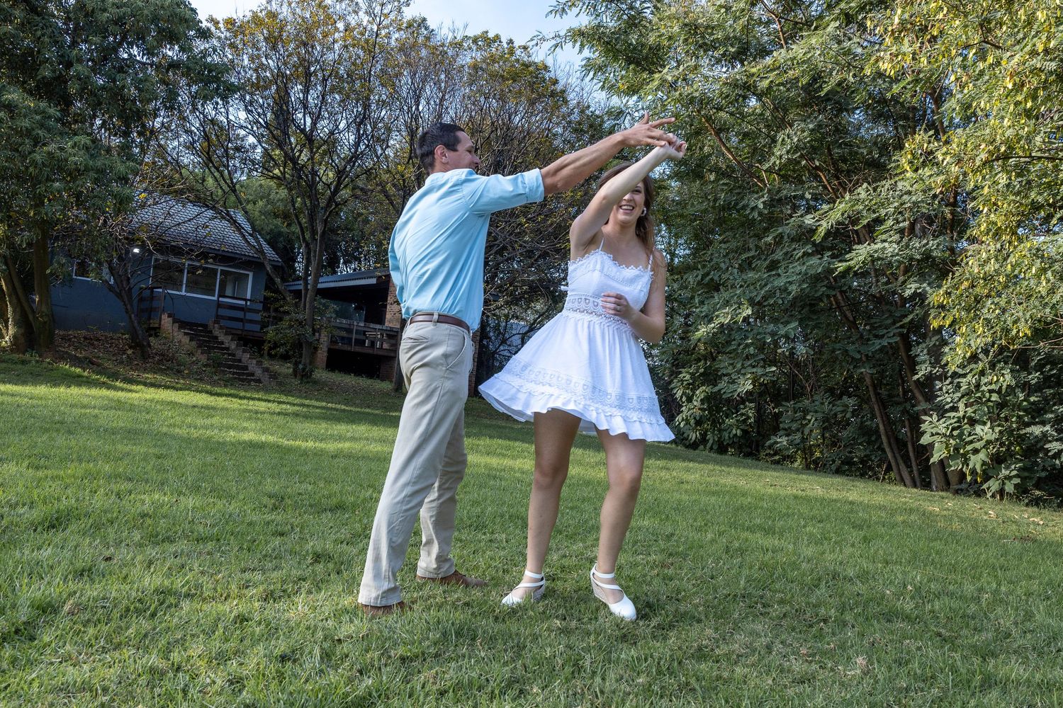 A couple dances joyfully on a green lawn surrounded by trees.