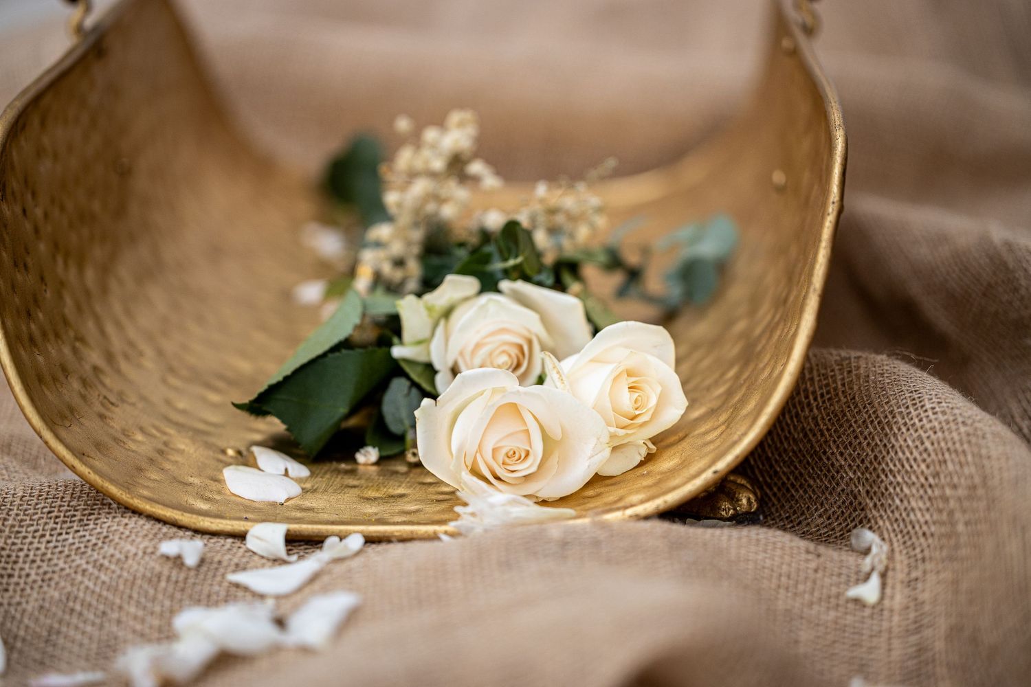 Elegant white roses and baby's breath arranged in a rustic gold bowl on burlap fabric.