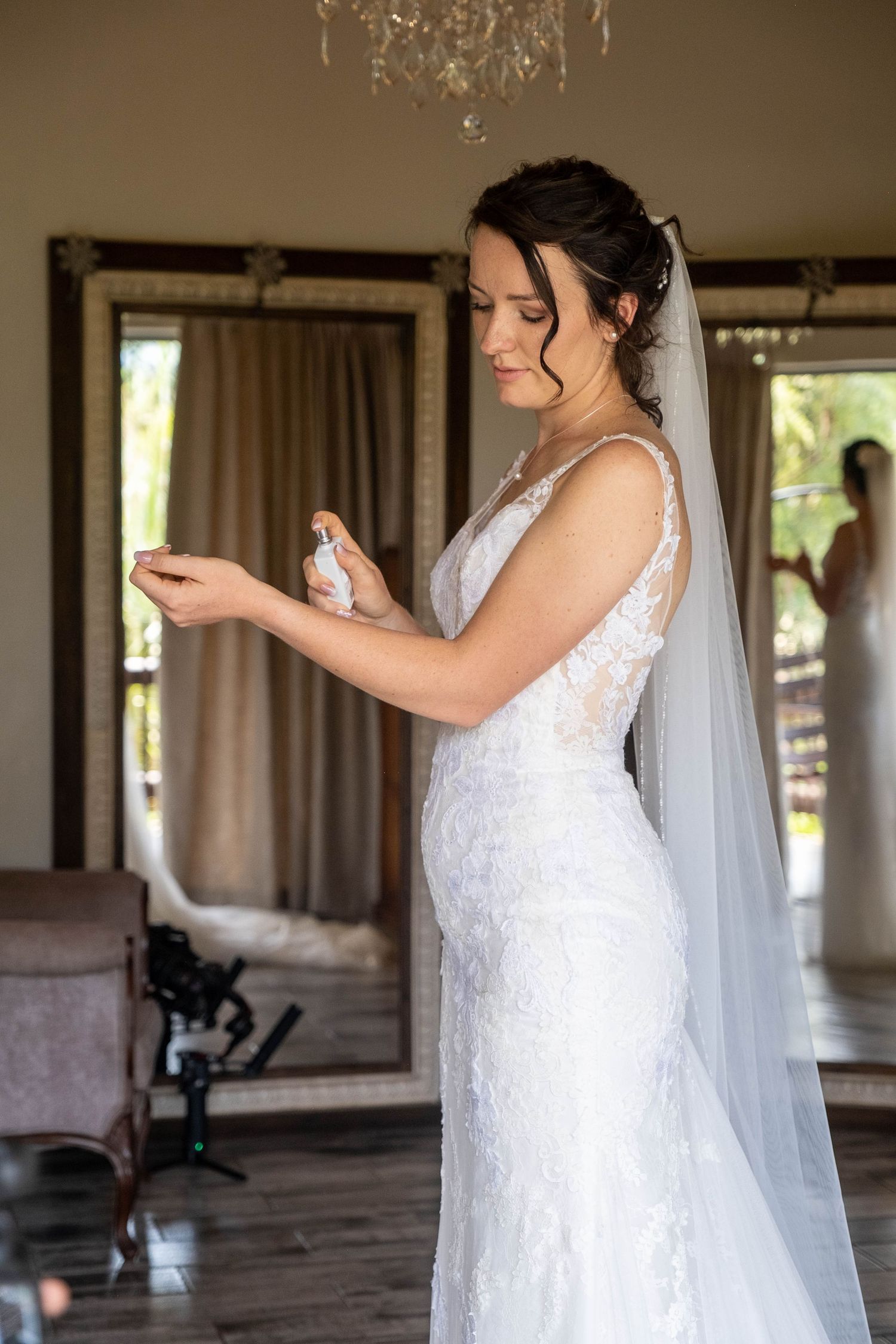 Elegant bride in a lace wedding gown and veil applying perfume while standing in front of a mirror.