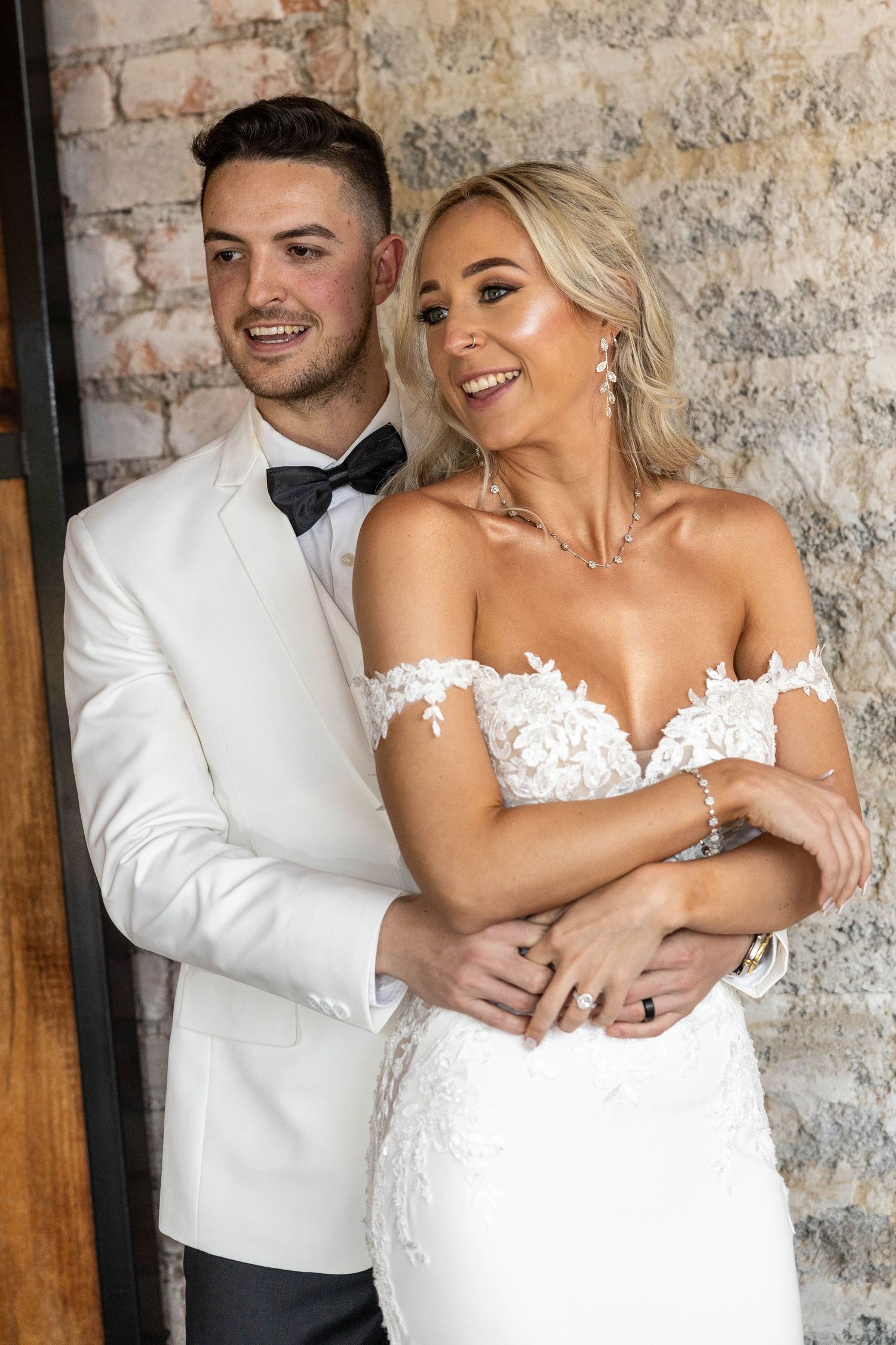 Smiling bride in lace off-shoulder gown and groom in white tuxedo posing together against stone wall.