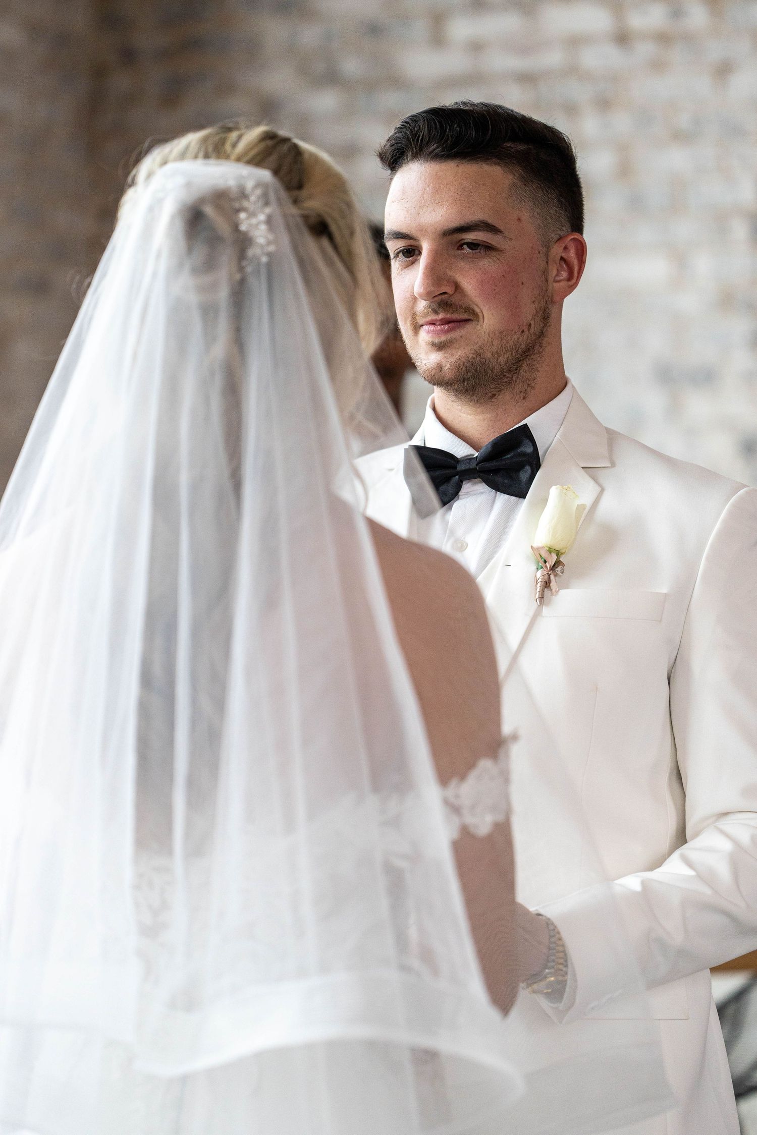 A groom in a white tuxedo gazes lovingly at his veiled bride during their intimate wedding ceremony.