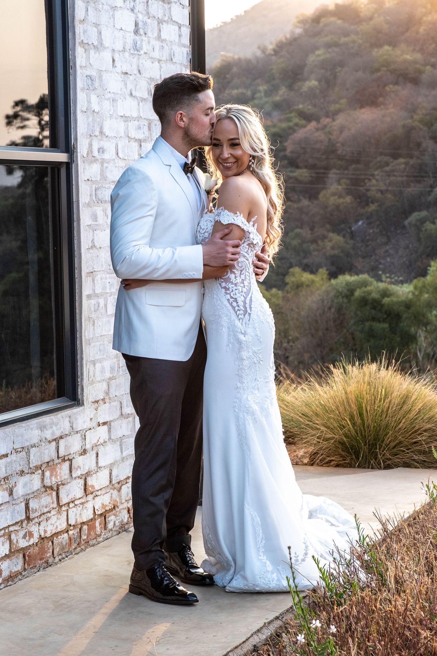 Groom in white blazer kisses bride in off-shoulder lace gown against brick wall with mountain backdrop.