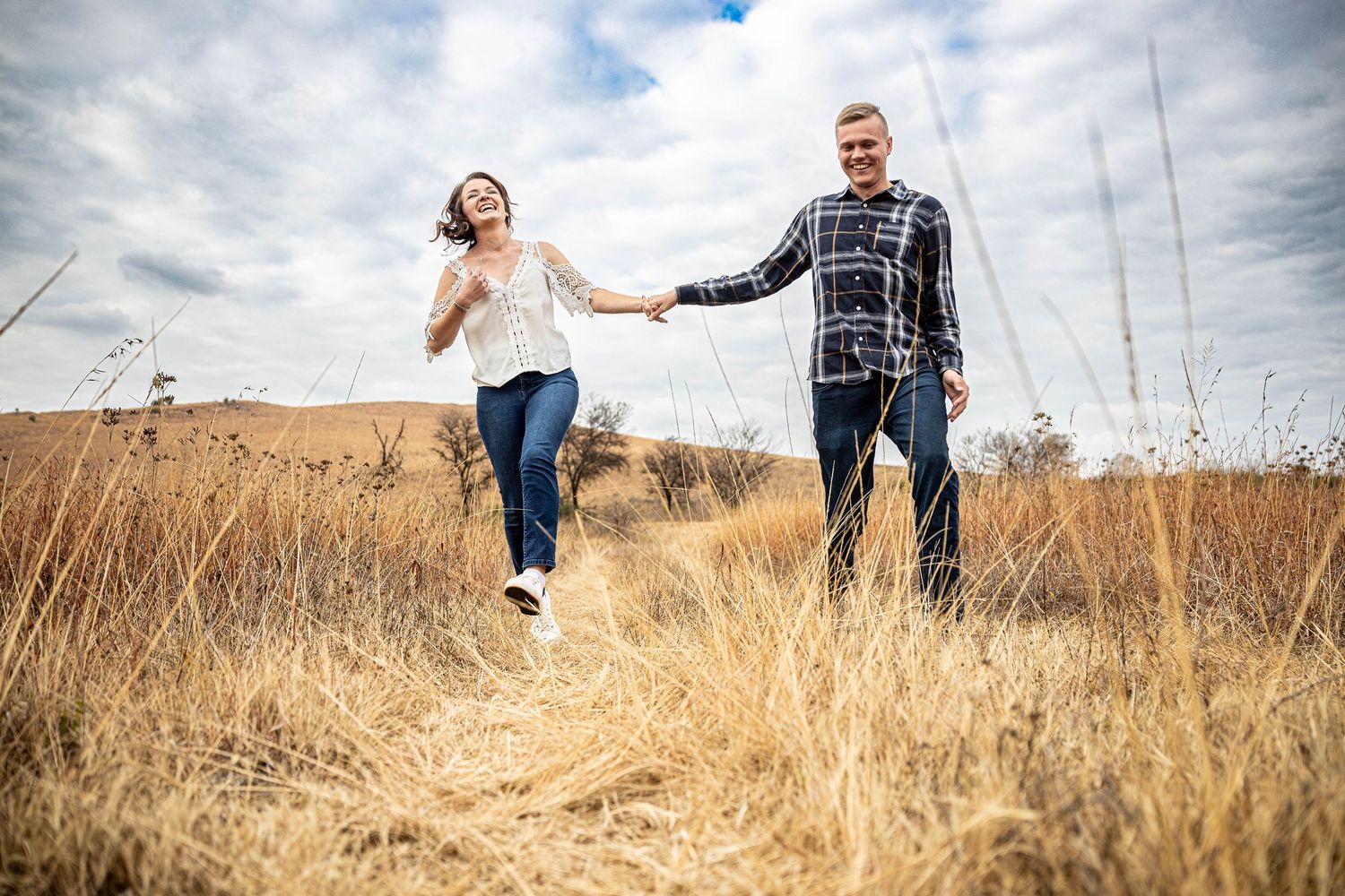 Joyful couple holding hands and walking through golden dry grass fields under a cloudy sky.