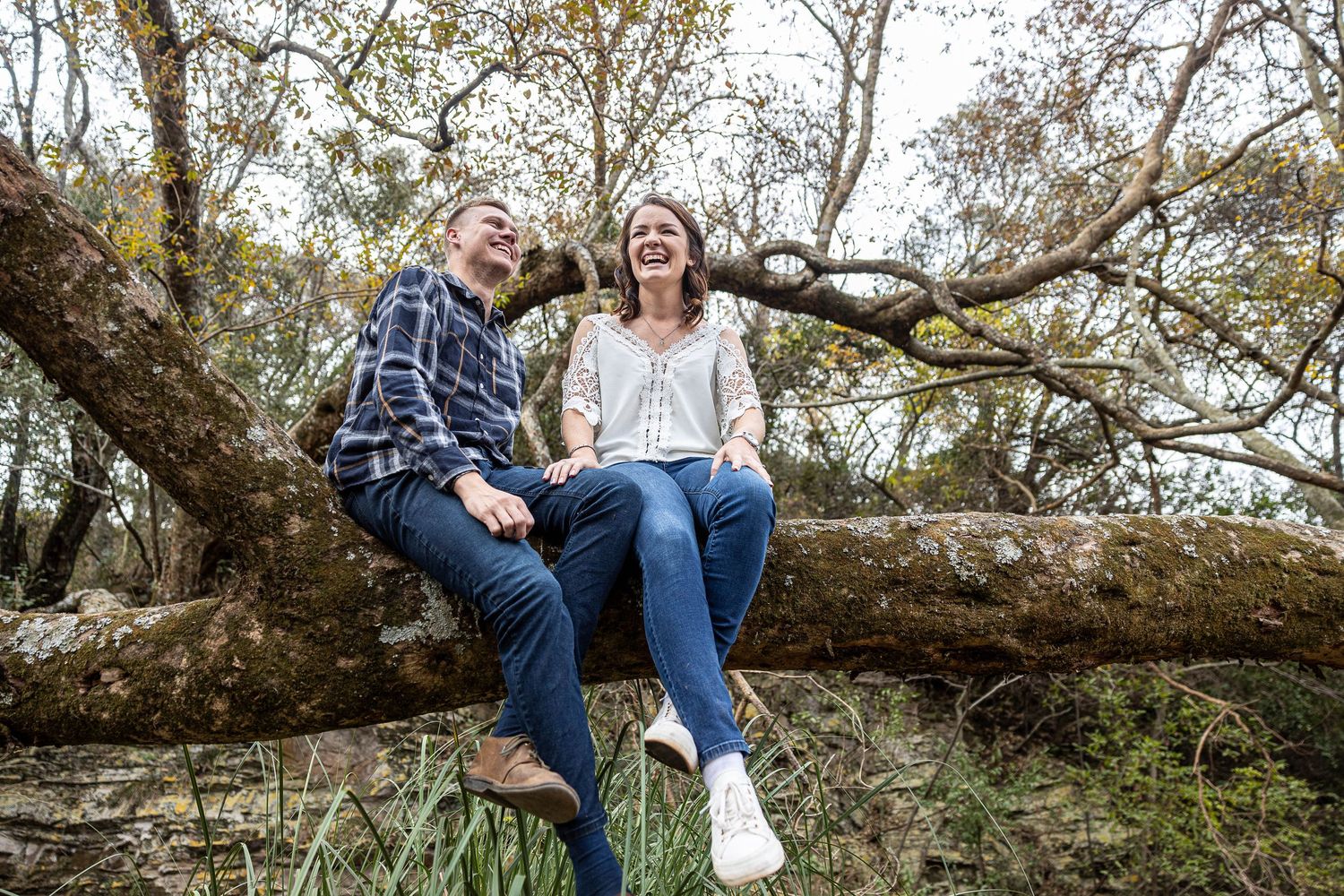 Happy couple laughs while sitting on fallen tree branch in wooded outdoor setting together.