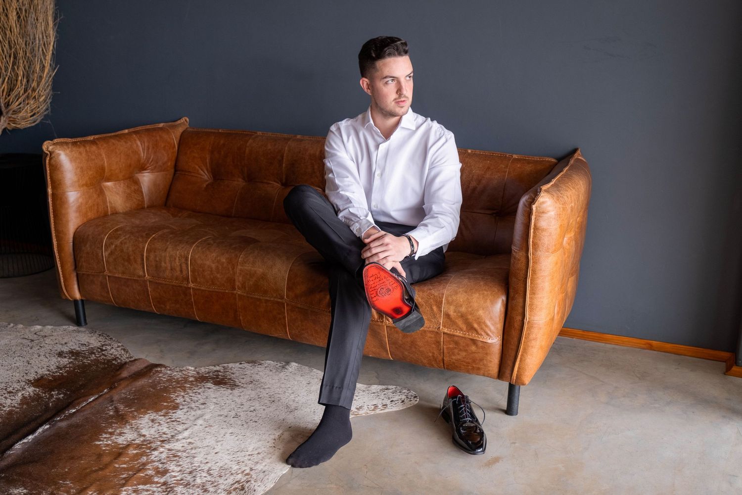Elegant man in white shirt sitting on brown leather sofa holding red dress shoe.