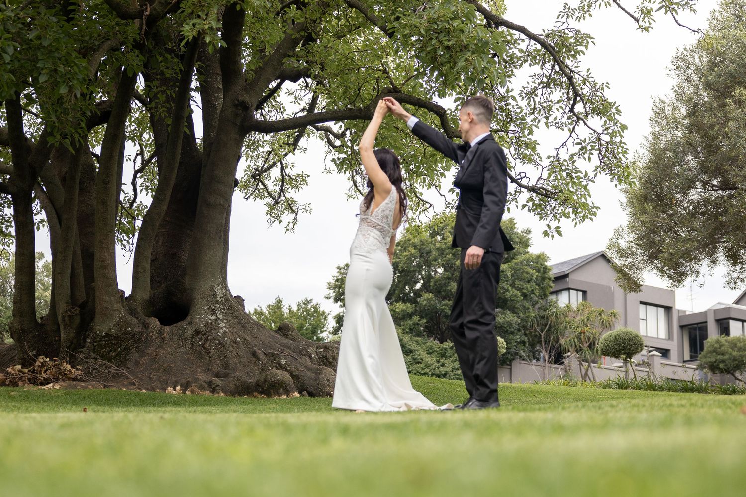 Bride and groom dancing outdoors on green lawn beneath large oak tree on their wedding day.