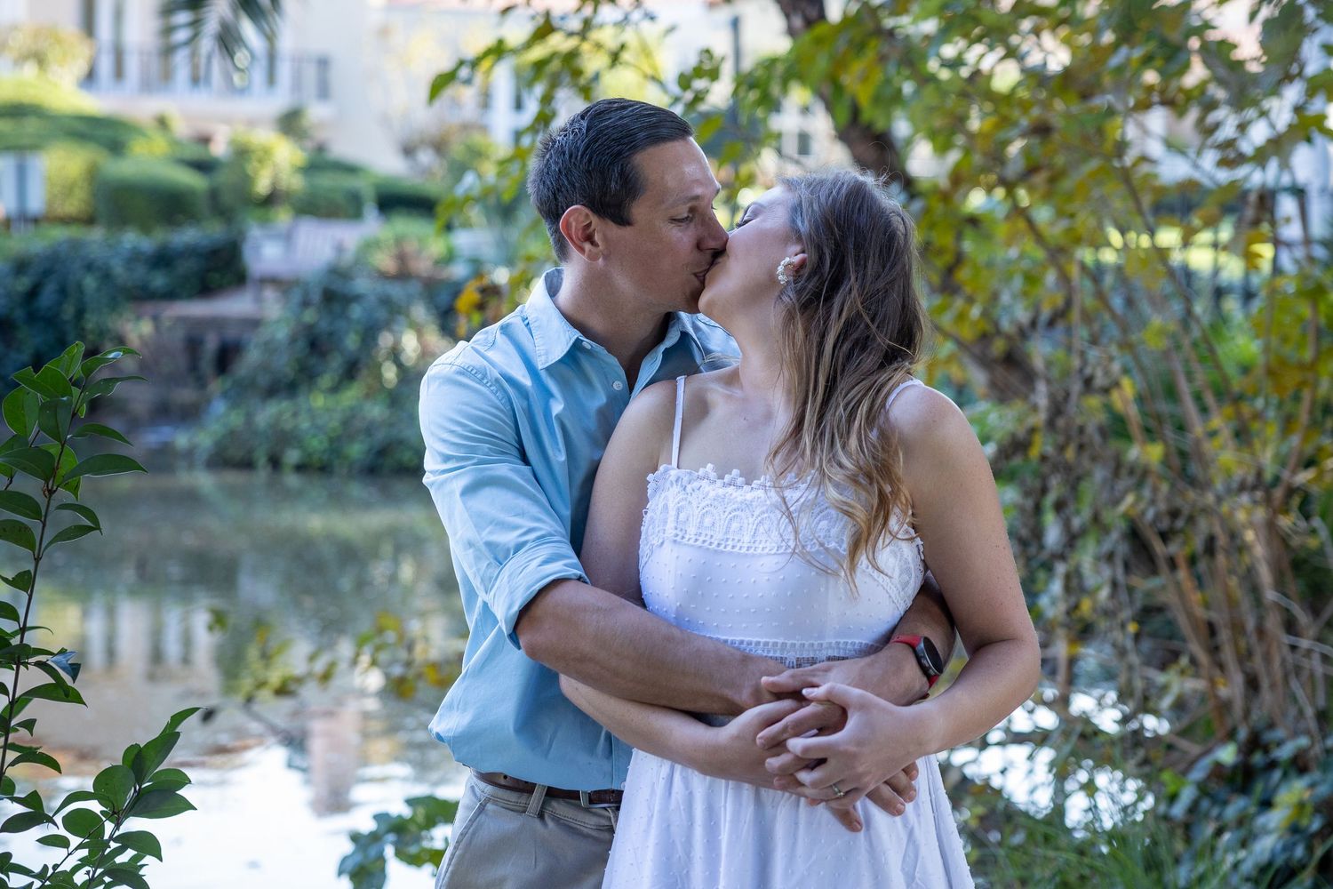 A couple shares a romantic kiss near a serene pond surrounded by lush greenery.