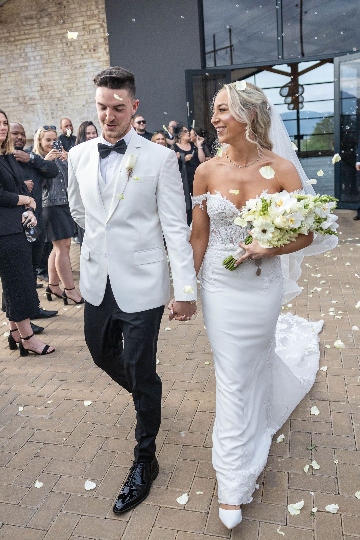 Newlyweds walk hand-in-hand through confetti shower, groom in white tuxedo jacket, bride in fitted lace gown holding bouquet.