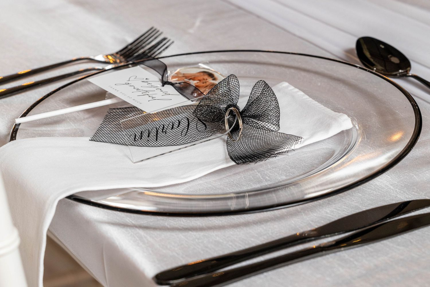 Elegant wedding place setting with clear charger plate, white napkin, black bow, and name card.
