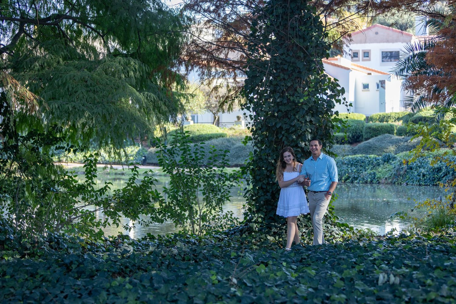 Couple embracing near an ivy-covered tree beside a tranquil pond in a scenic garden park.