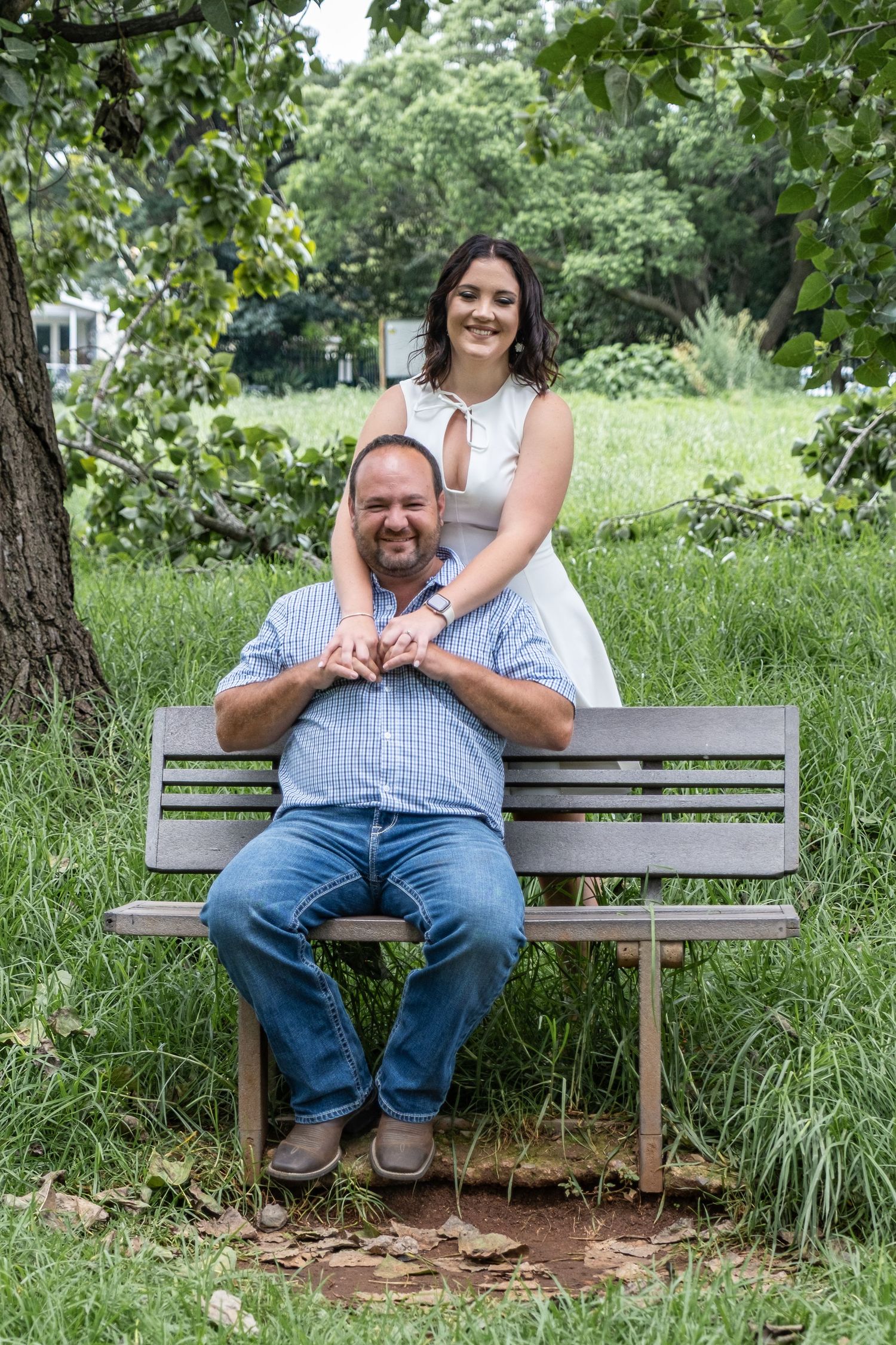 A woman in a white dress stands behind a man seated on a park bench, both smiling warmly.
