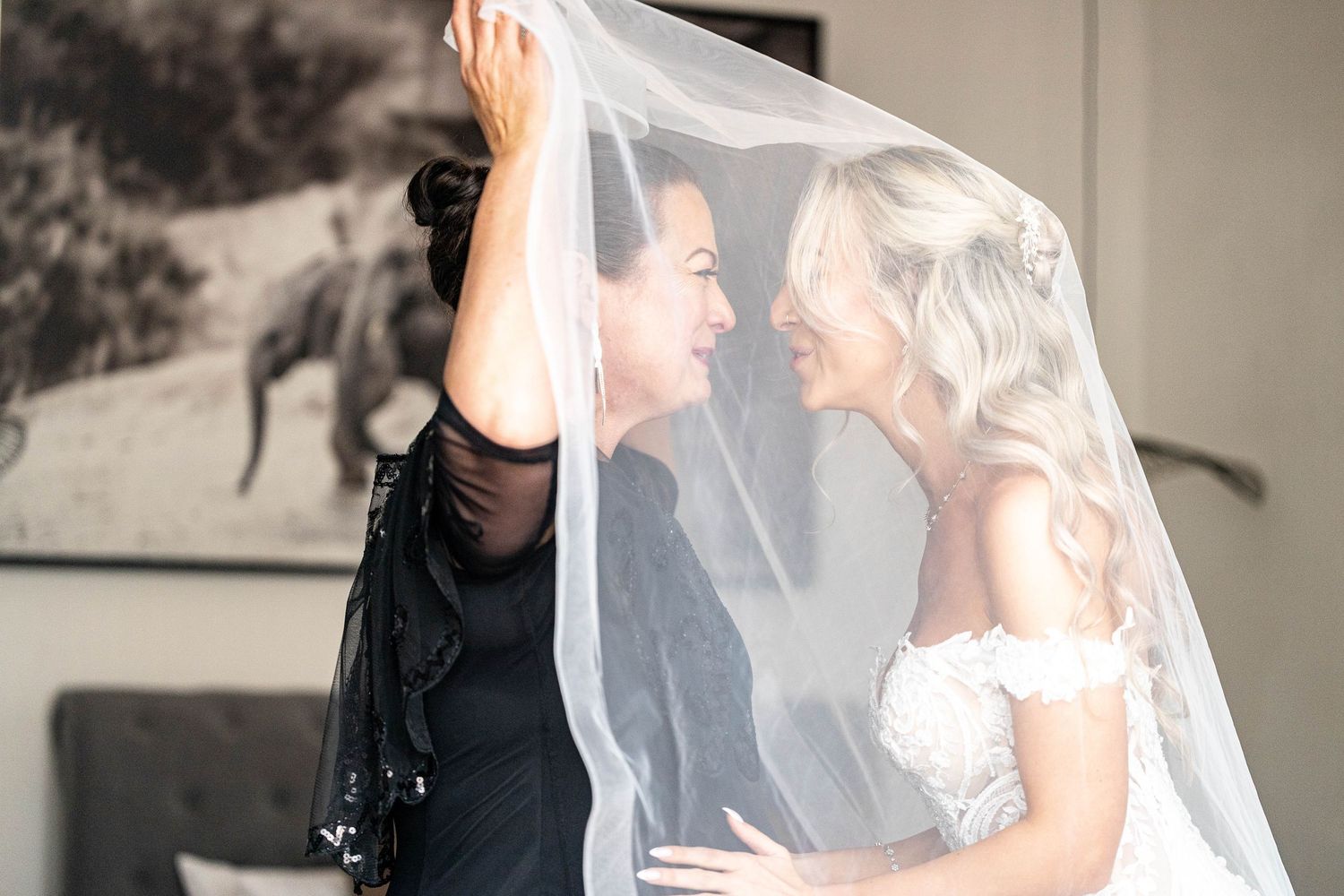 Bride and guest share an intimate moment under wedding veil while photographer captures the scene.