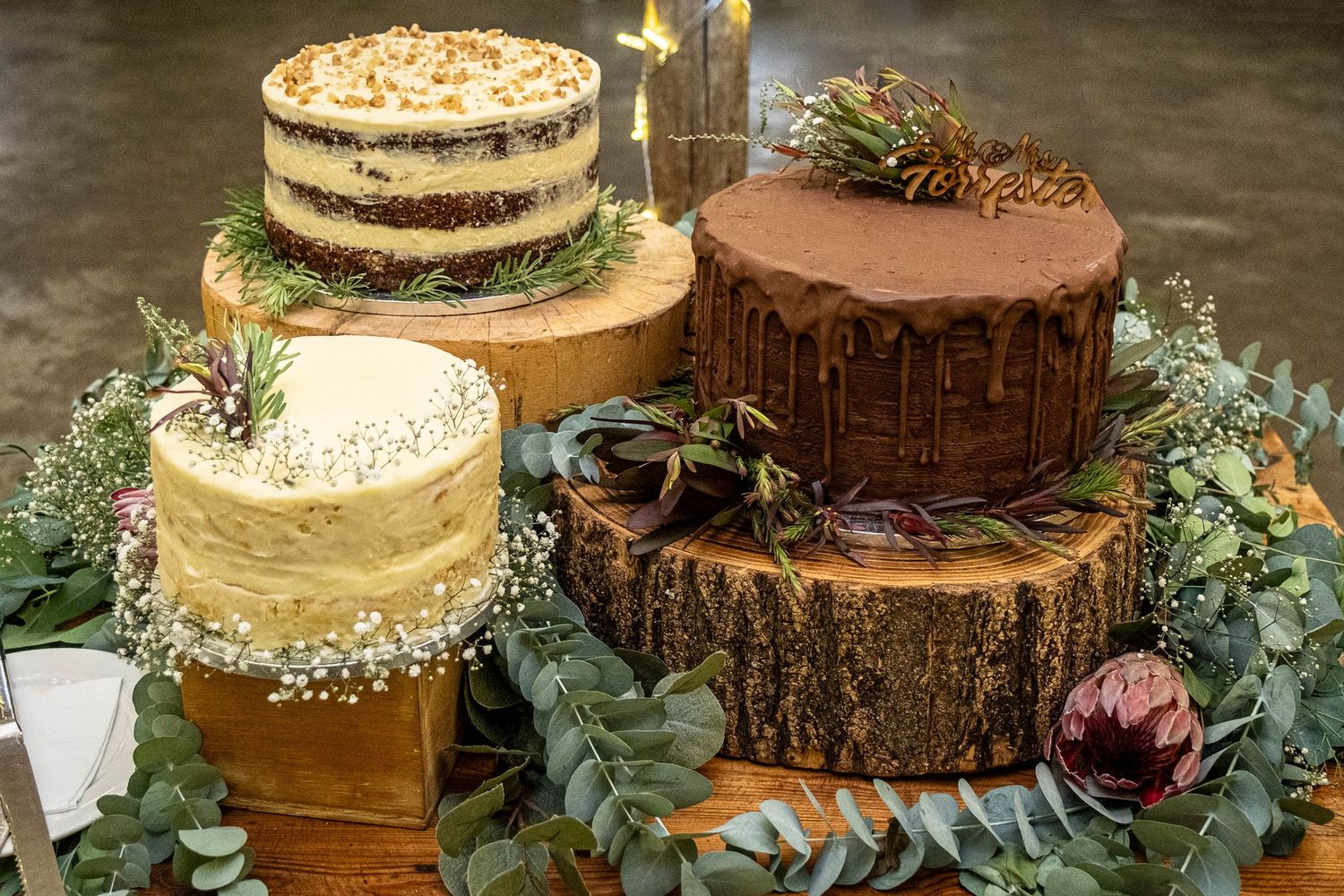 Four elegant wedding cakes displayed on rustic wooden slices surrounded by lush greenery and flowers.