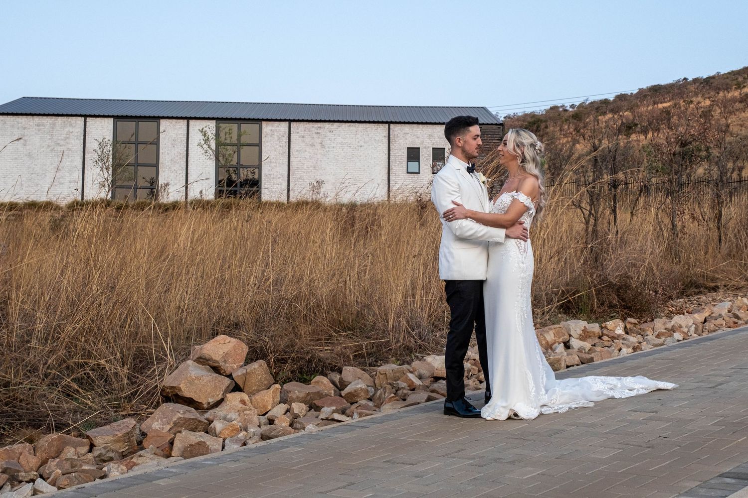 Bride and groom share a tender embrace on a pathway surrounded by golden dry grass and rocky landscape.