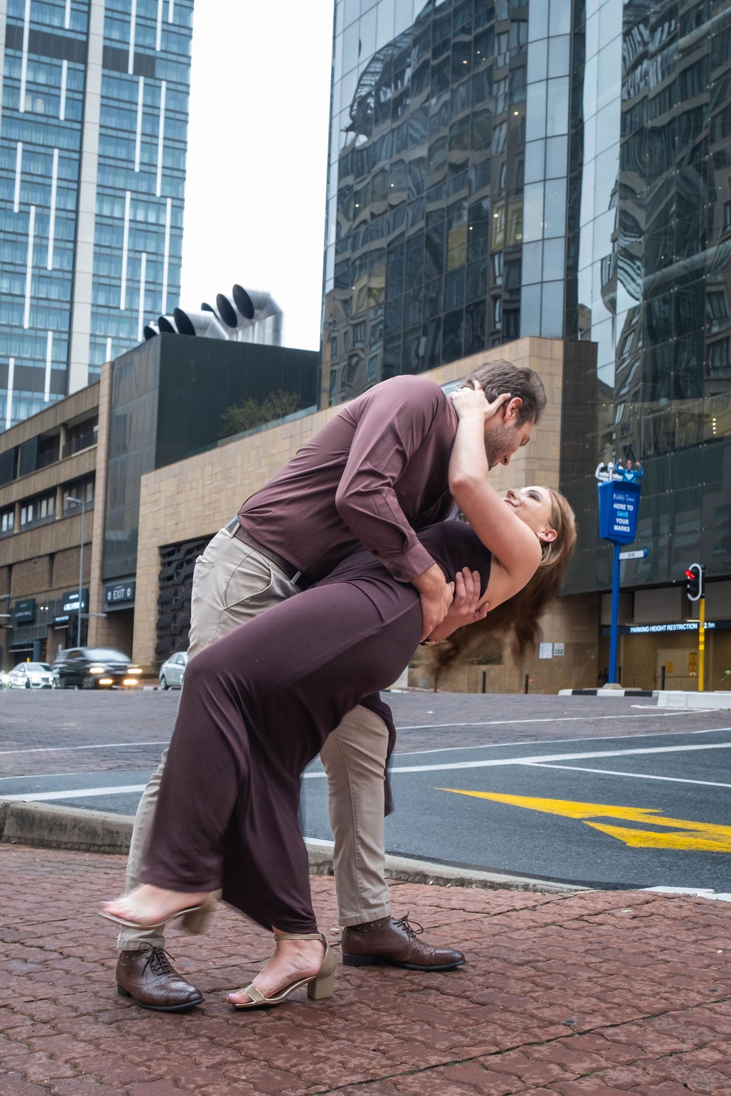 Couple shares passionate dip kiss on urban brick sidewalk surrounded by glass office building reflections.