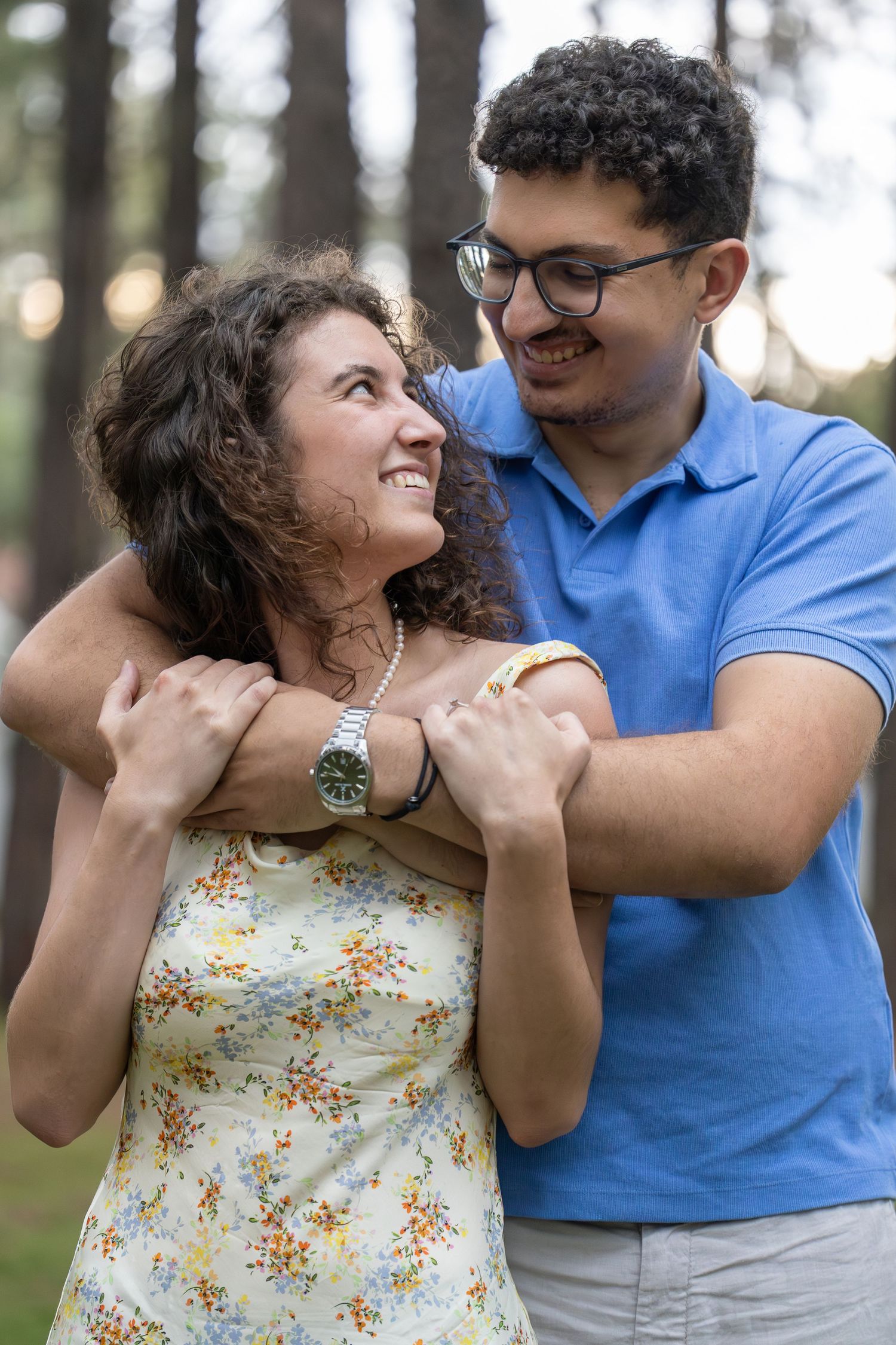 A man in a blue polo hugs a curly-haired woman in a floral dress from behind in a forest setting.