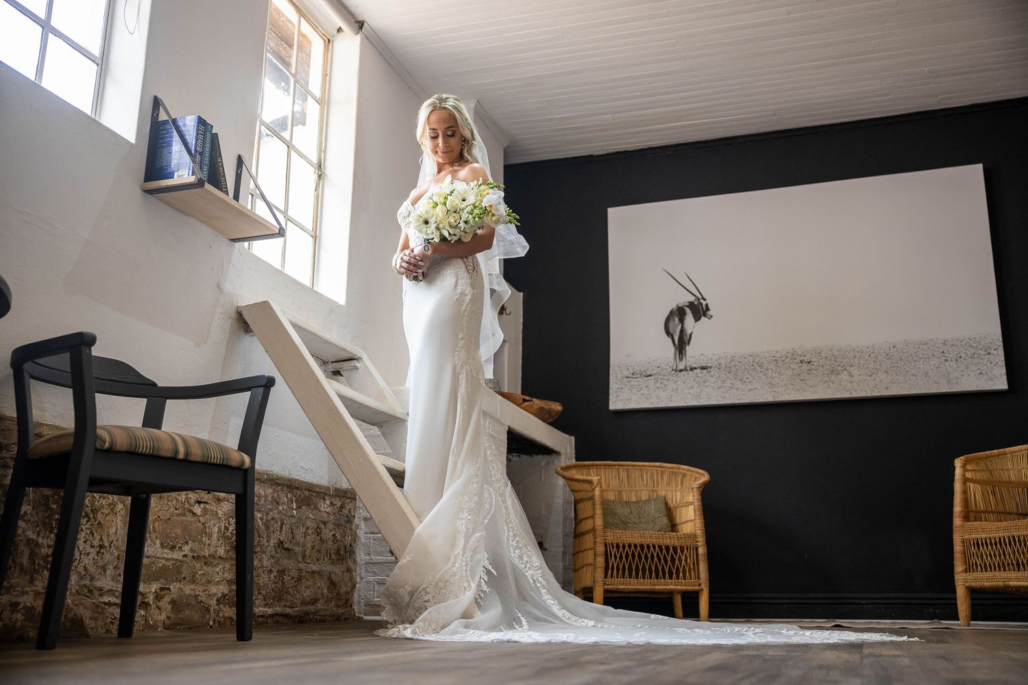 Full-length view of bride in lace mermaid gown with long train holding white bouquet indoors.