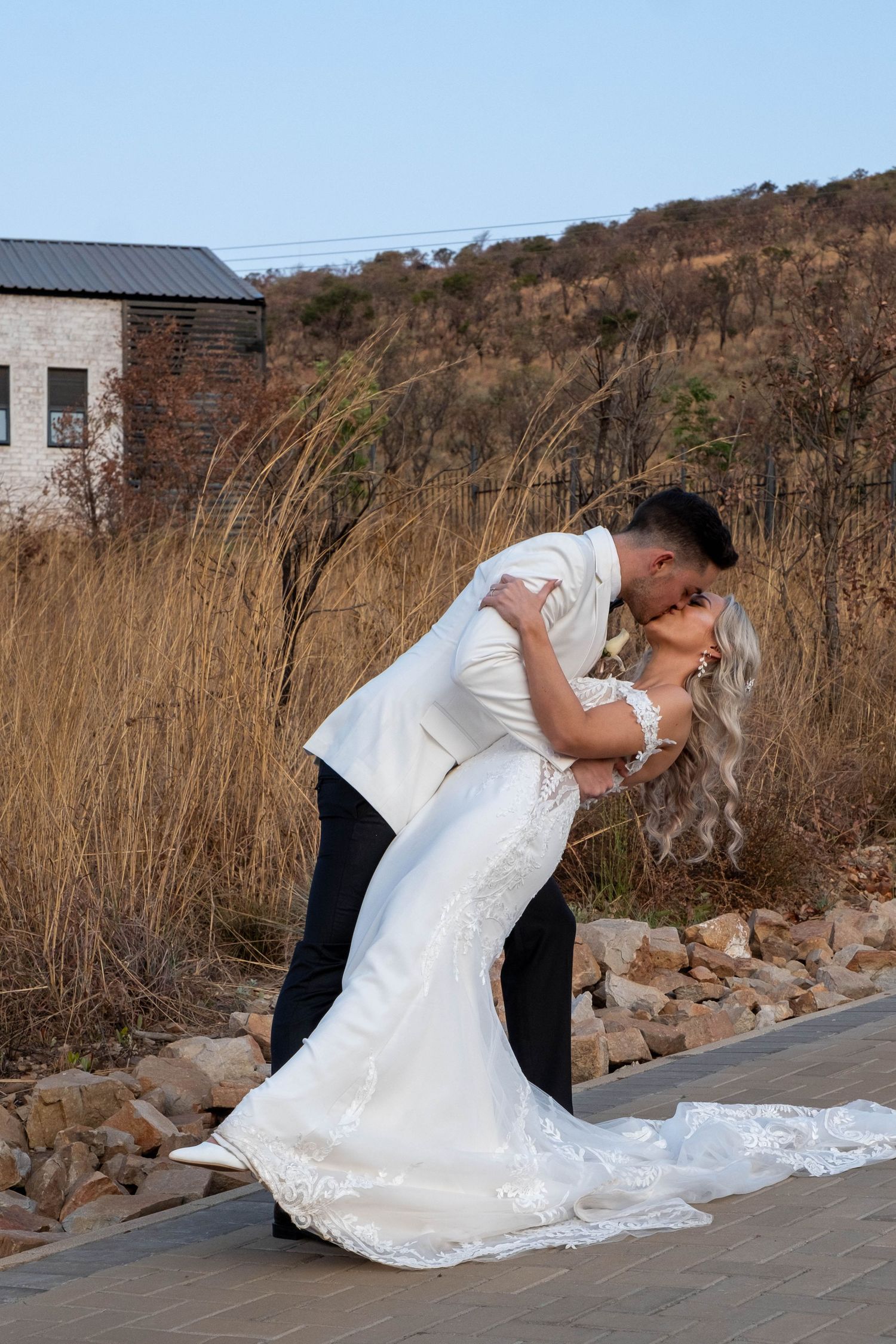 Groom dips bride for a romantic kiss outdoors with golden dry grass and rocky hillside in background.
