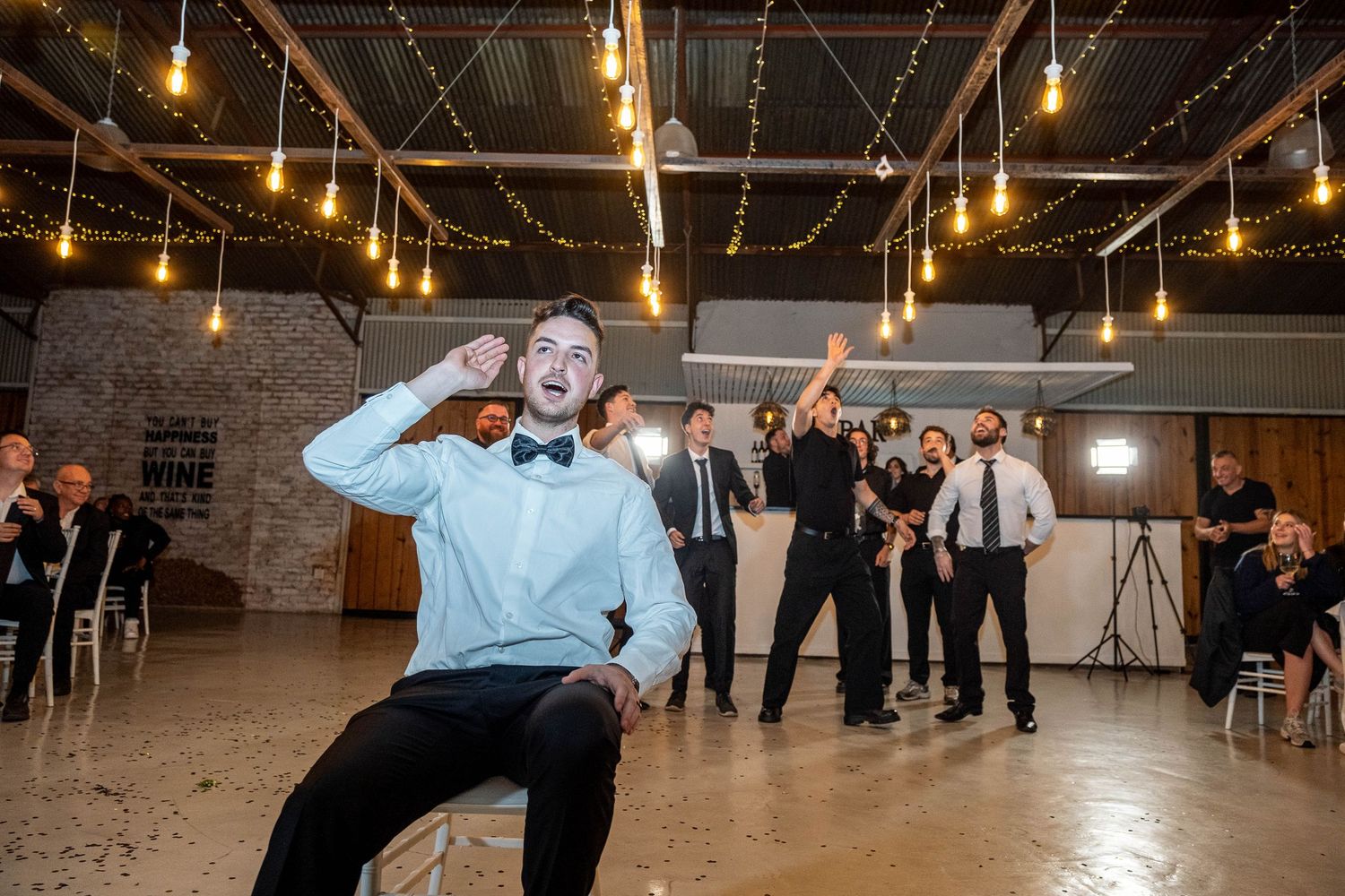 Groom sitting in chair tossing garter to group of men at wedding reception with Edison lights.